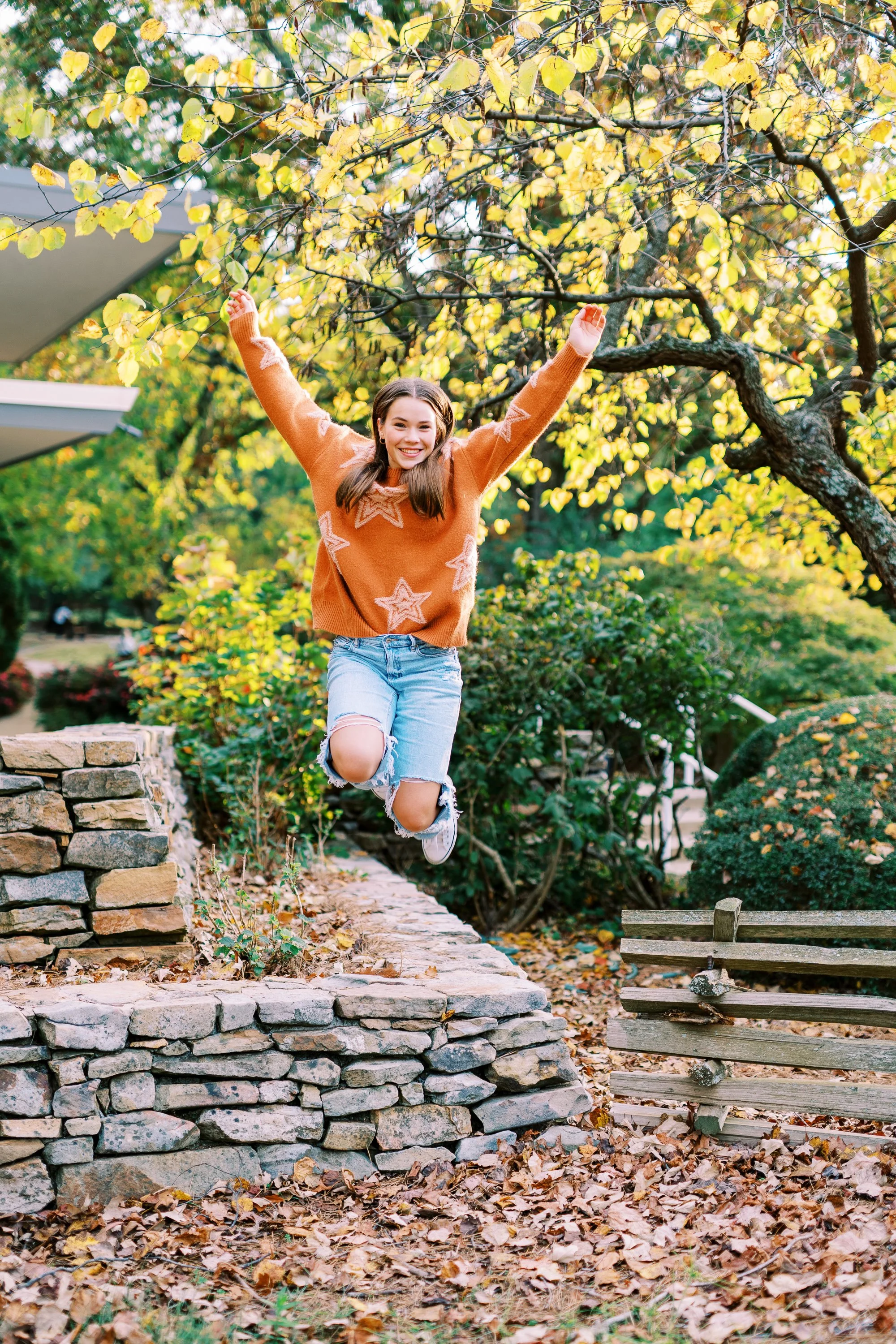 Senior portrait of Hannah jumping playfully on a stone wall in an orange sweater at Kennesaw Mountain Battlefield Park in Kennesaw, Georgia