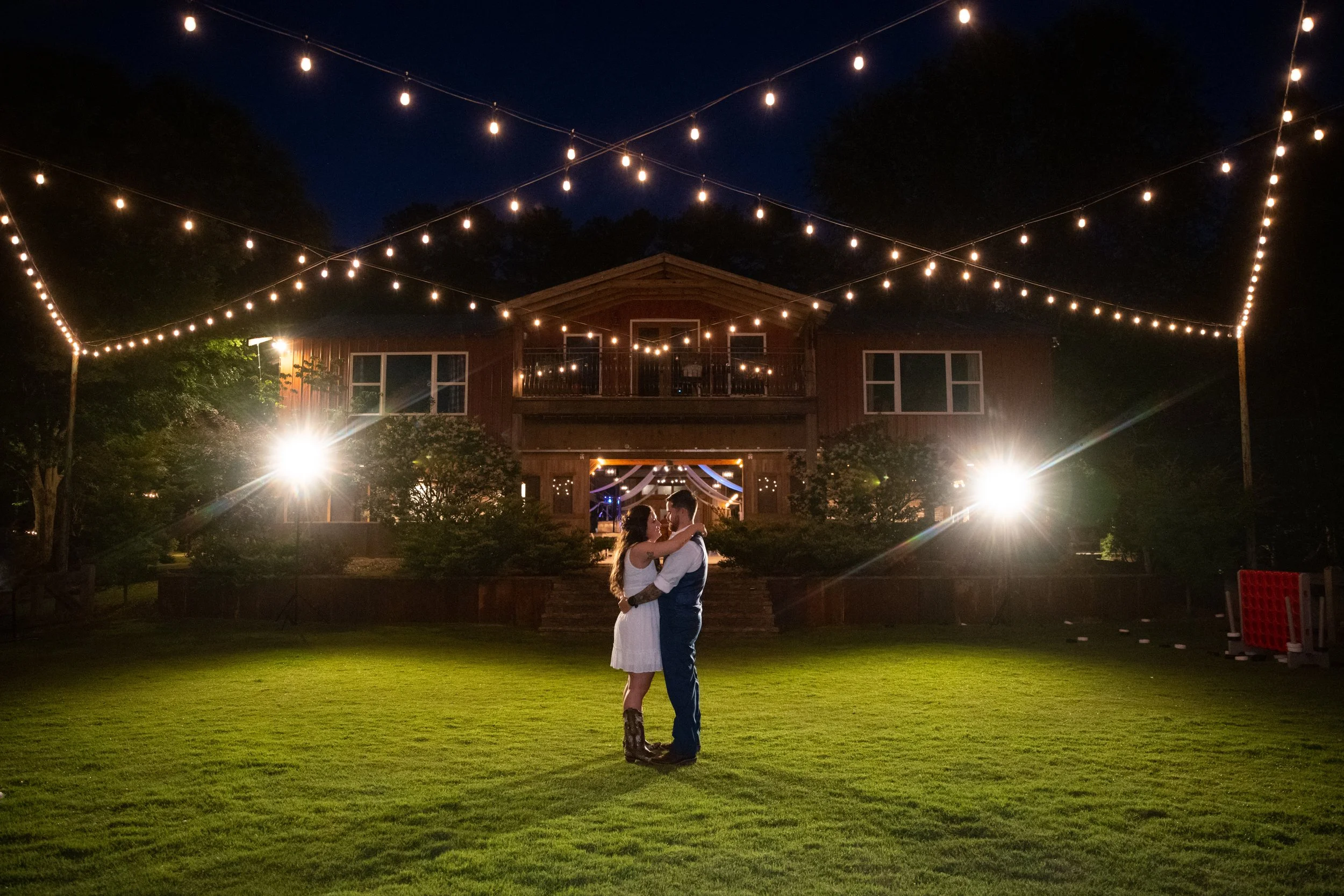Wide nighttime view of the couple embracing on the lawn with the venue lit by string lights