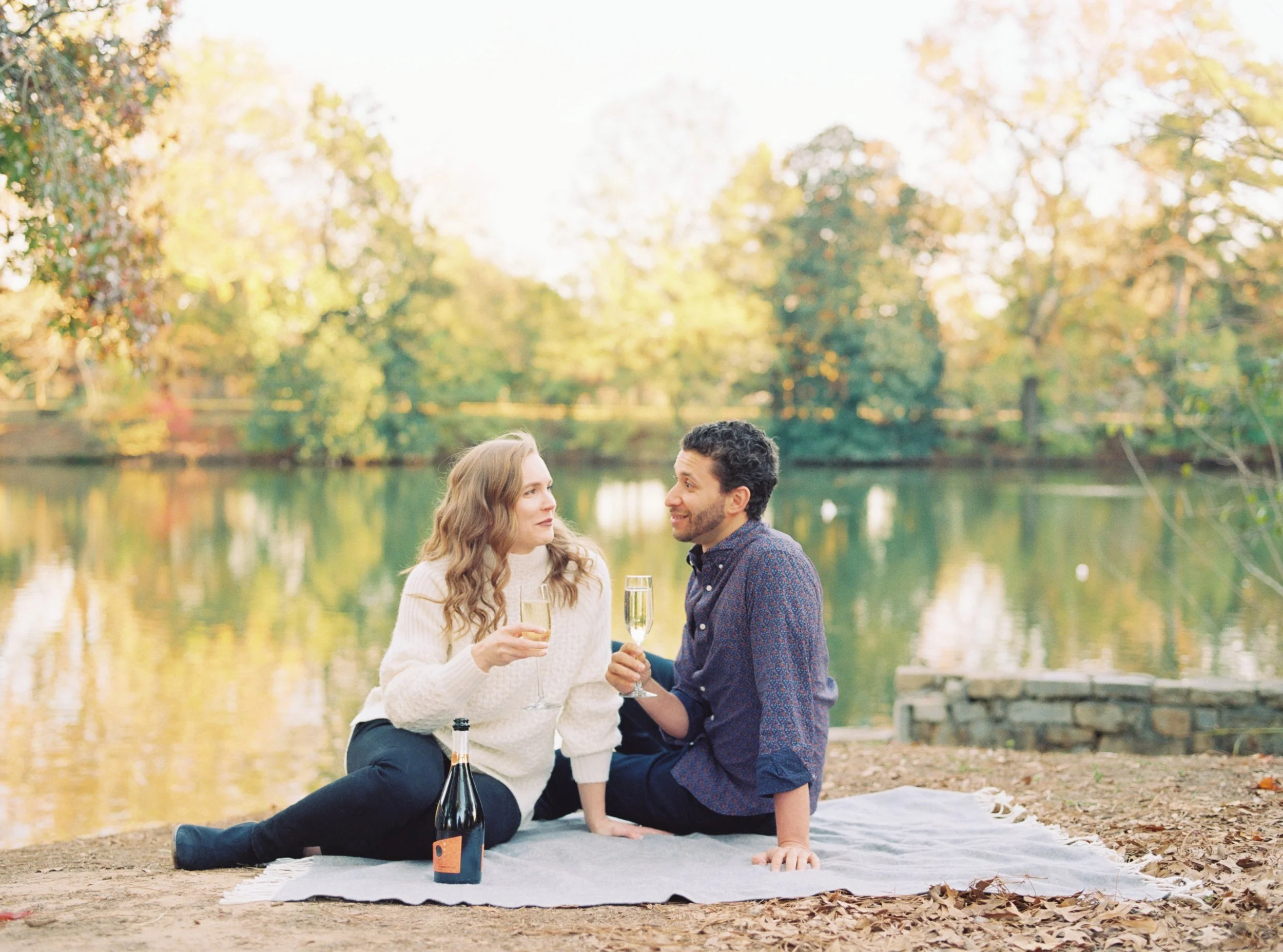 Couple sitting together on a blanket by the lake, sharing a toast at golden hour
