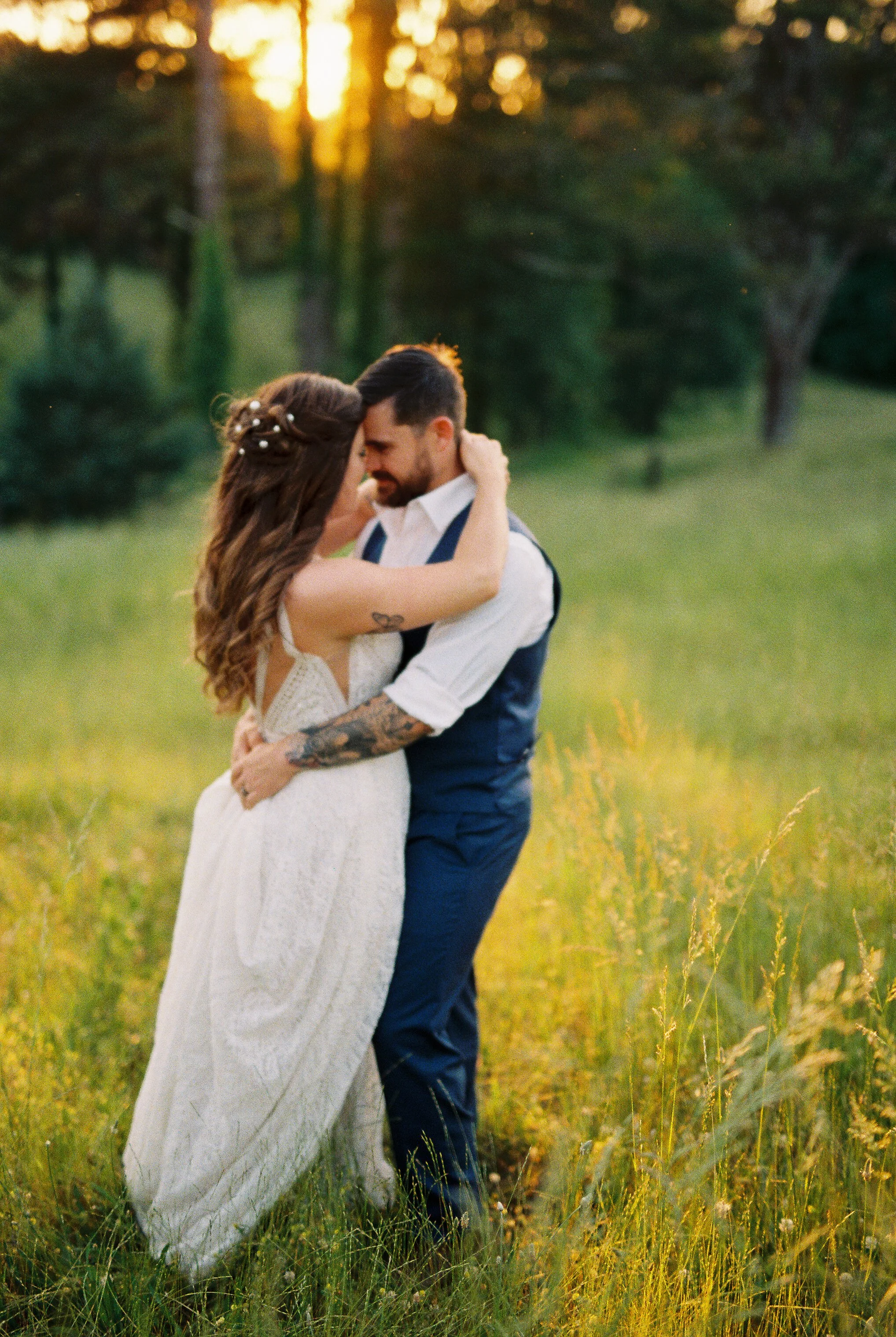 Bride and groom embracing closely in a sunlit meadow at golden hour, sun setting through the trees behind them