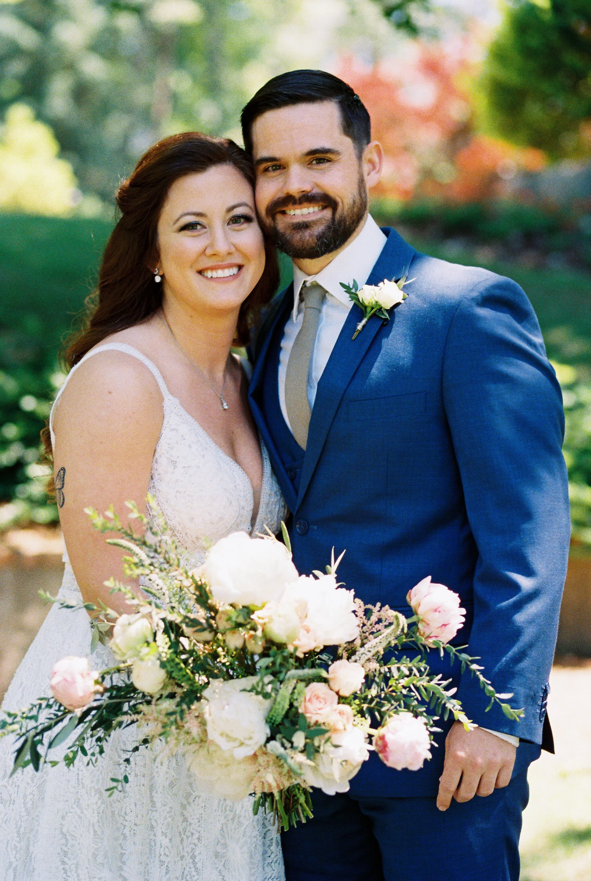Bride and groom smiling directly at the camera in a close embrace outdoors, bride holding a white and greenery bouquet
