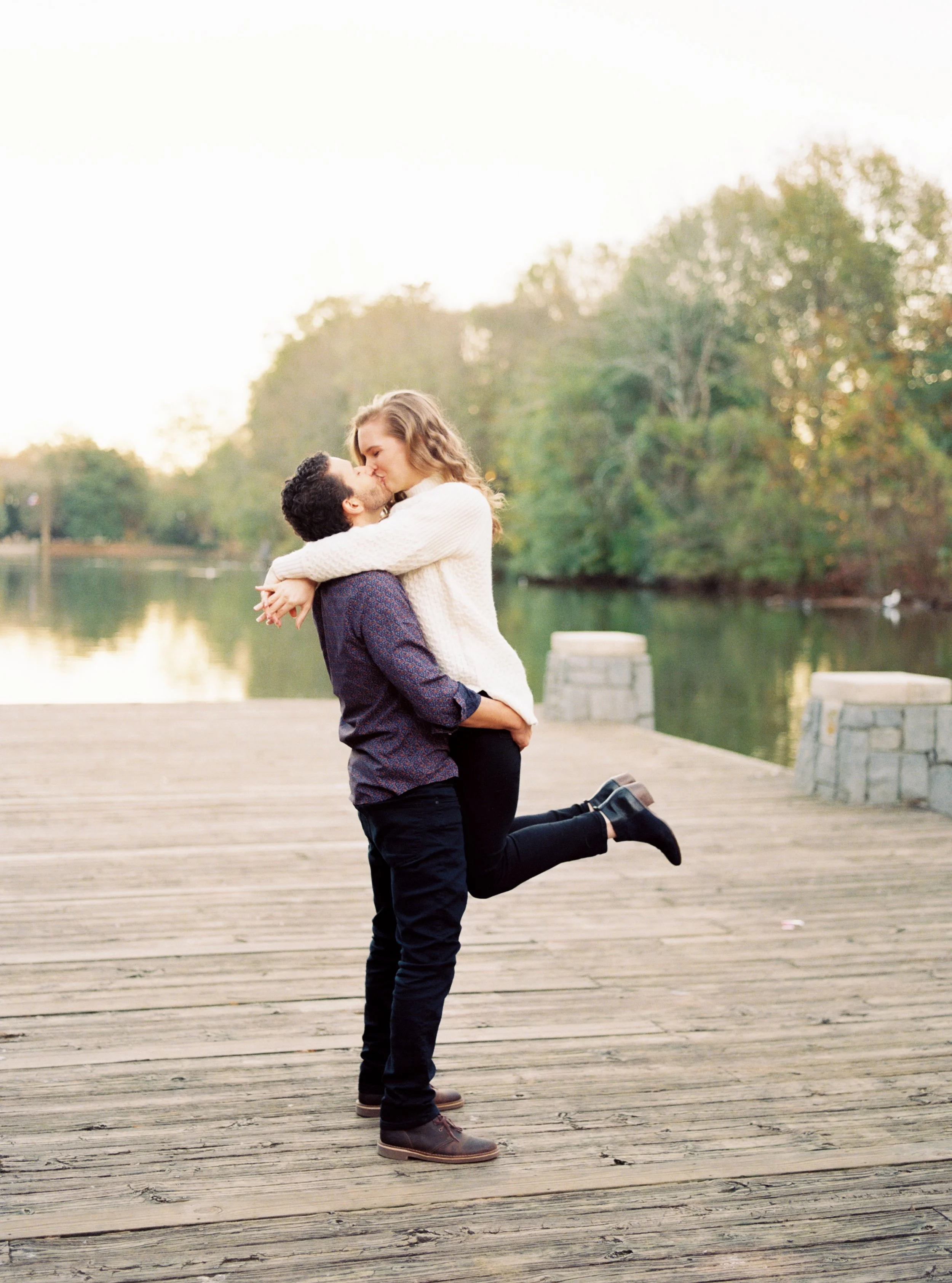 Full-body shot of man lifting woman with her leg kicked back on the wooden dock, lake and trees in the background