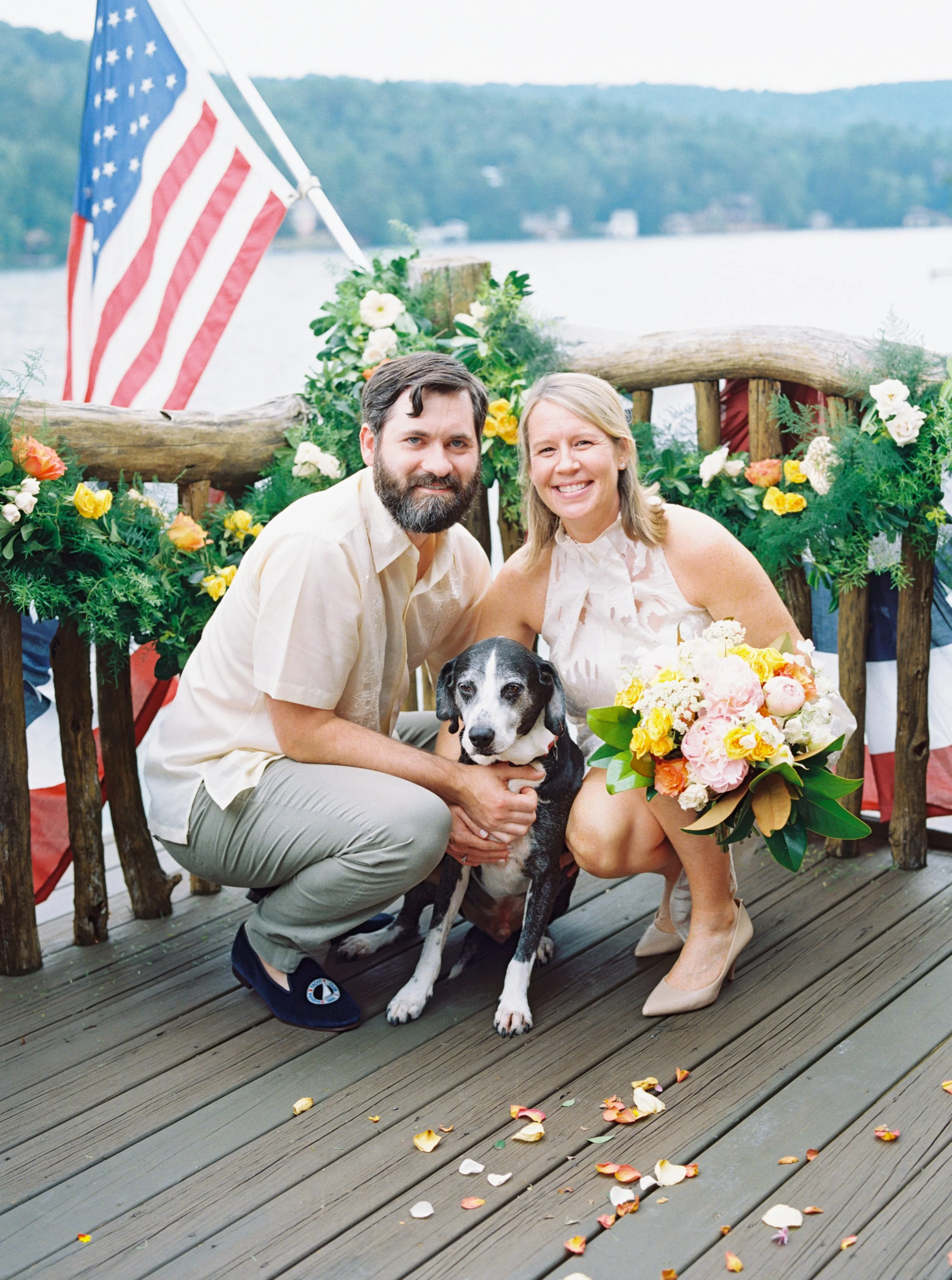 Bride and groom crouched down with their dog on the deck, American flag and lake in the background