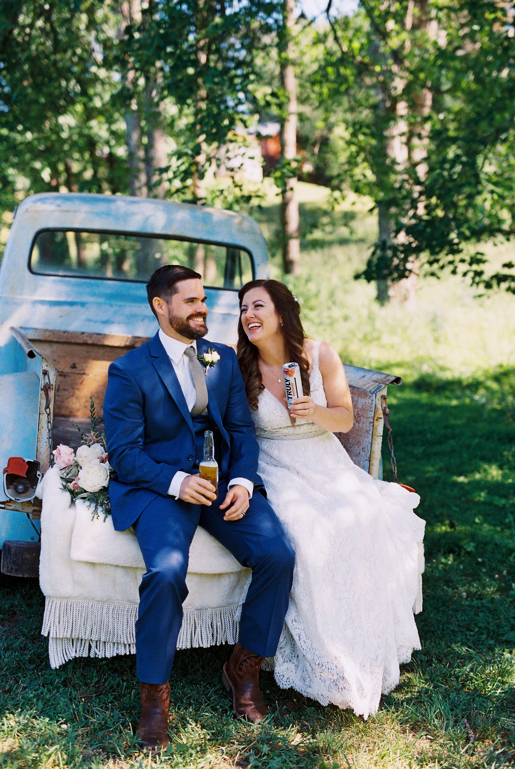 Bride and groom laughing together while holding drinks, seated on the vintage truck's tailgate