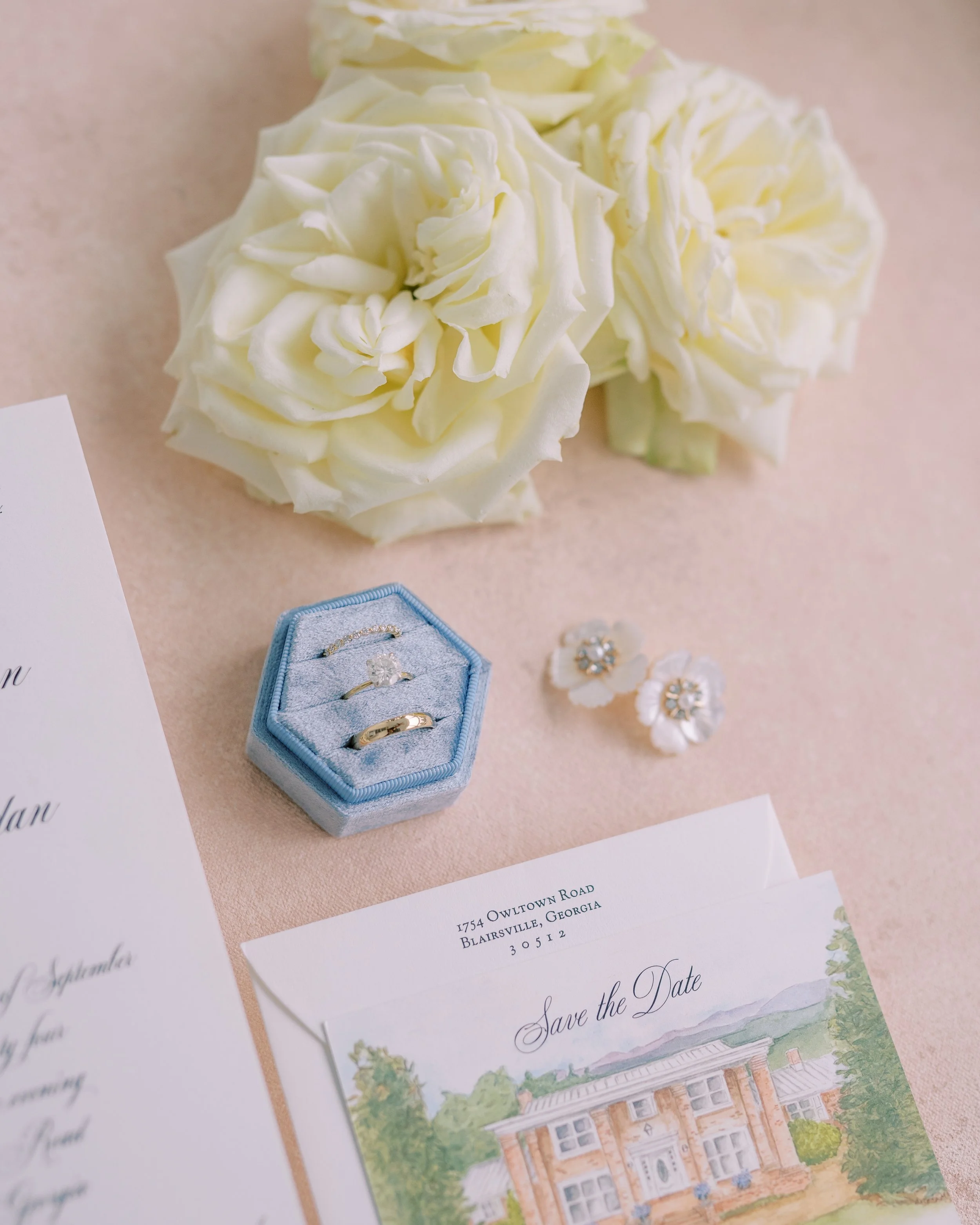 Flat lay of wedding invitation suite, rings, earrings, and ivory roses