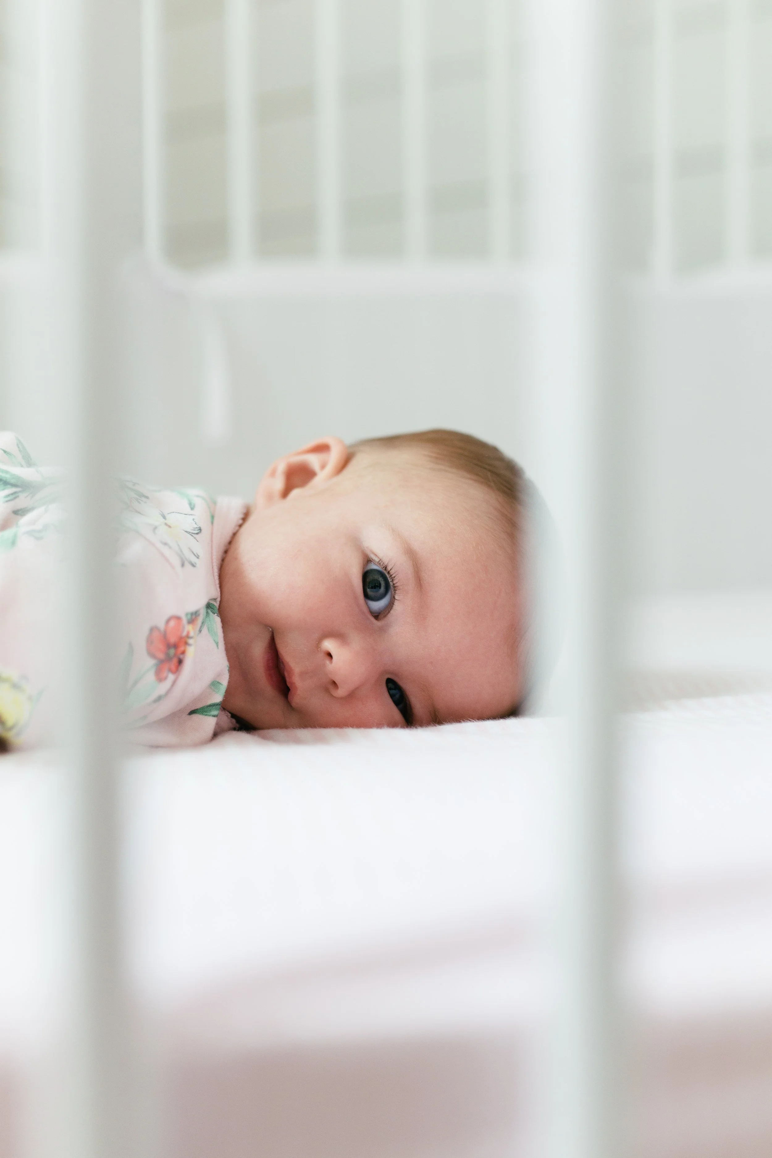 A baby lying down on a white surface inside a crib, looking through the crib bars with blue eyes, wearing a floral-patterned outfit.