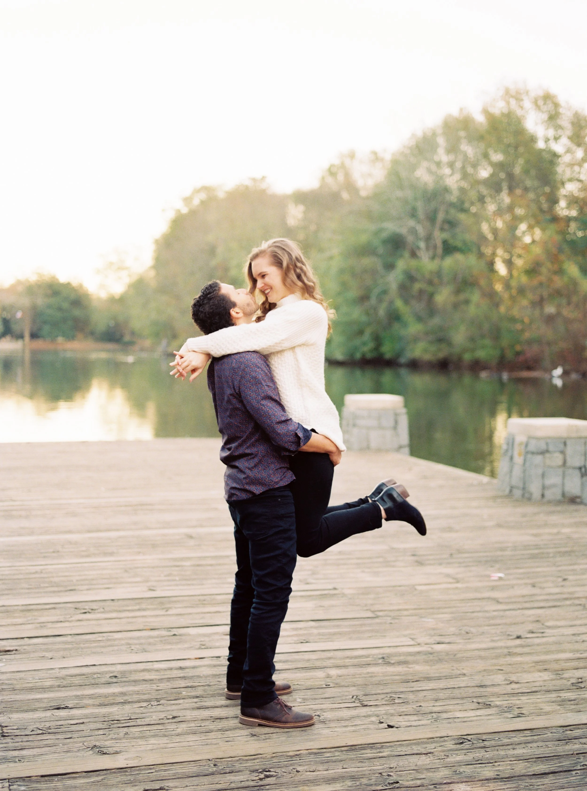 Man joyfully lifting woman with her leg kicked back on a wooden dock overlooking the lake