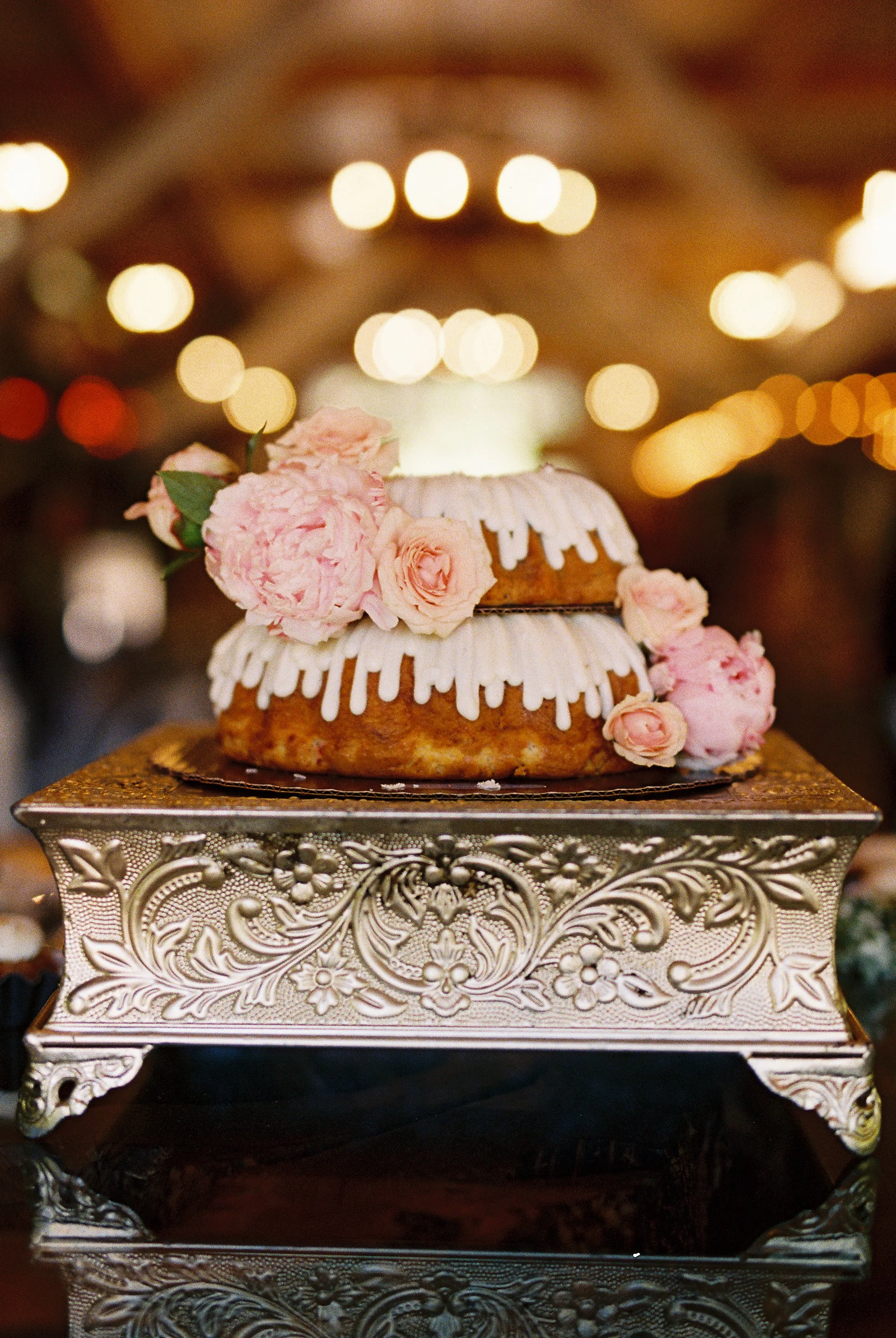 Two-tier bundt-style wedding cake with white icing drizzle and pink roses, displayed on an ornate silver stand with string lights glowing in the background