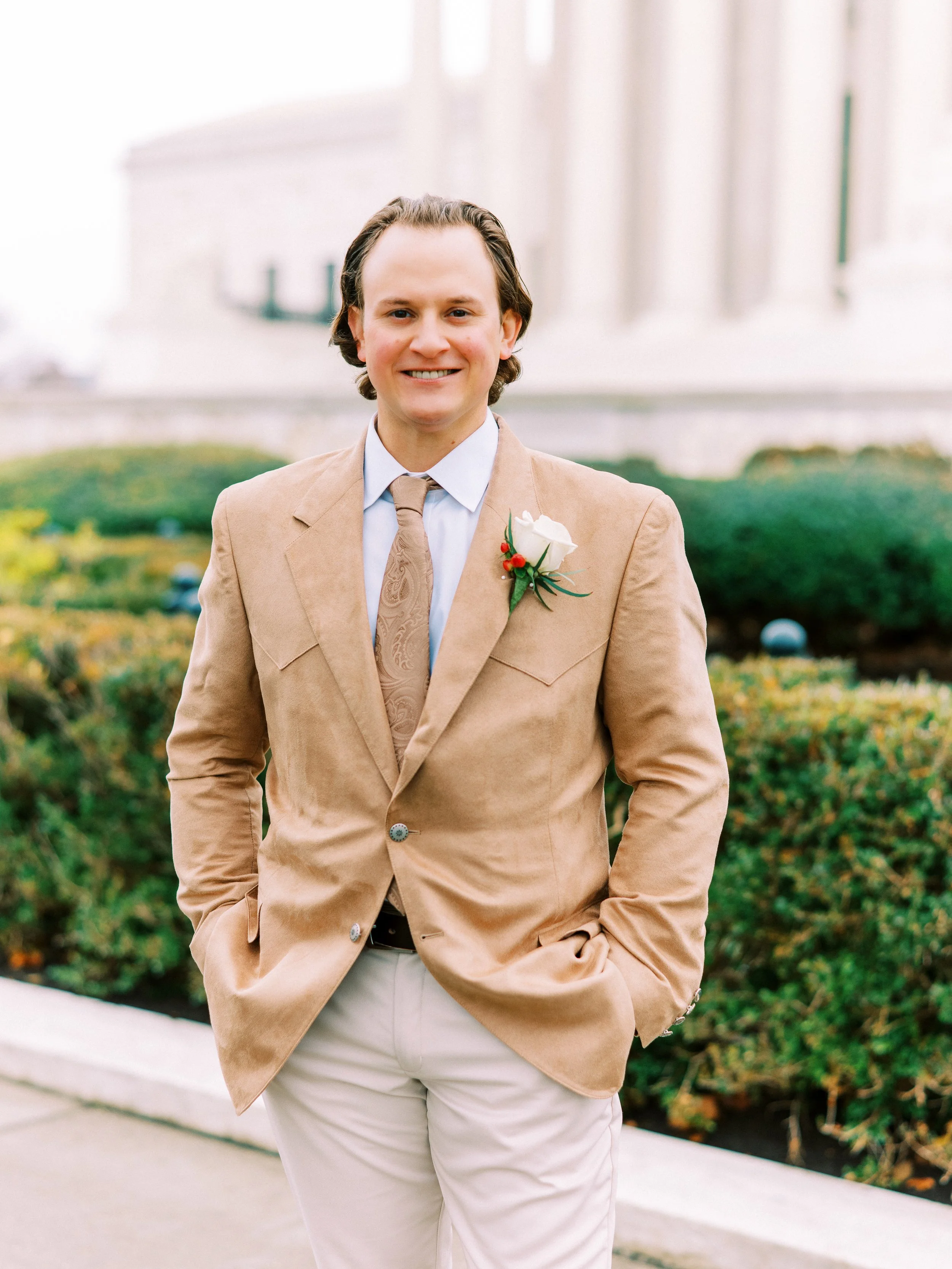 Groom standing solo in a garden with hedges behind him