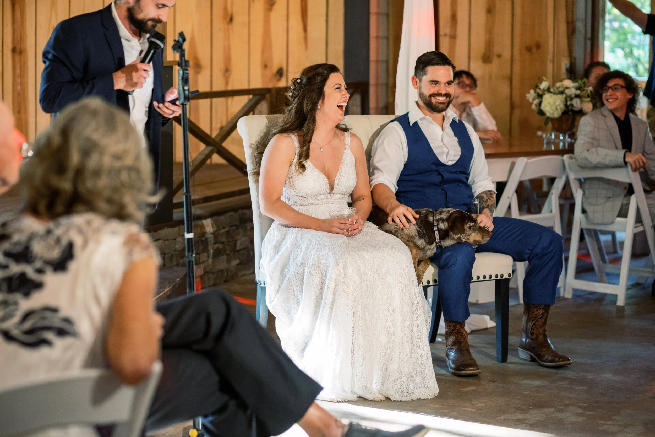 Bride and groom seated together while a guest speaks at the microphone