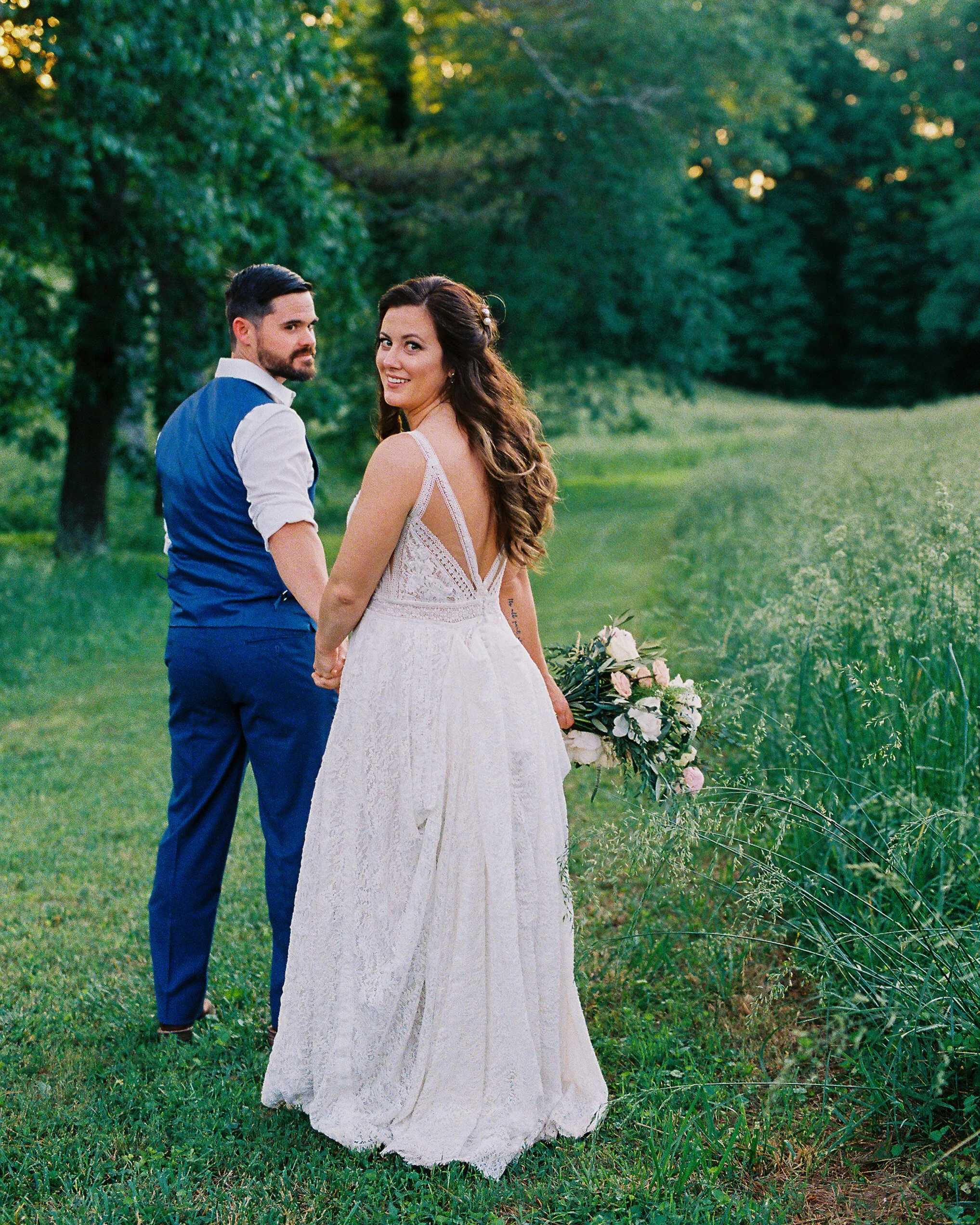 Bride looking back over her shoulder with a smile while walking hand in hand with the groom through a grassy field, her backless lace gown and bouquet visible