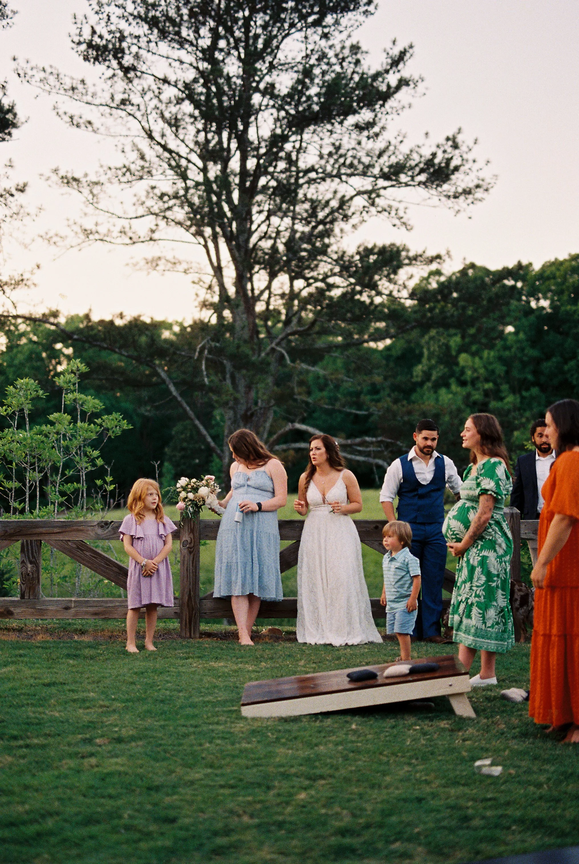 Bride and family members watching children play cornhole together on the lawn at dusk