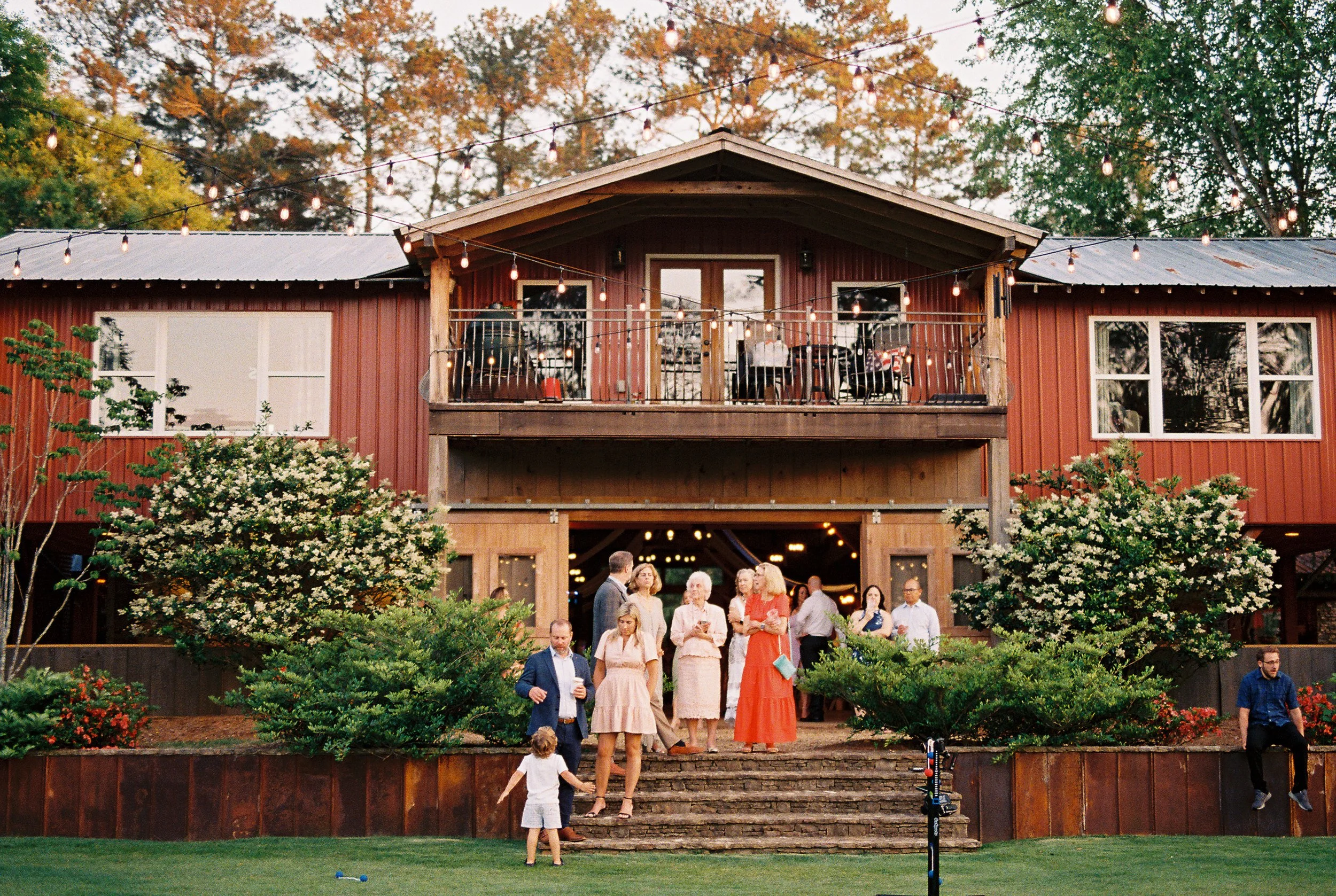 Wide exterior view of the red barn venue with its two-story balcony strung with lights, guests gathered on the steps and a child running across the lawn