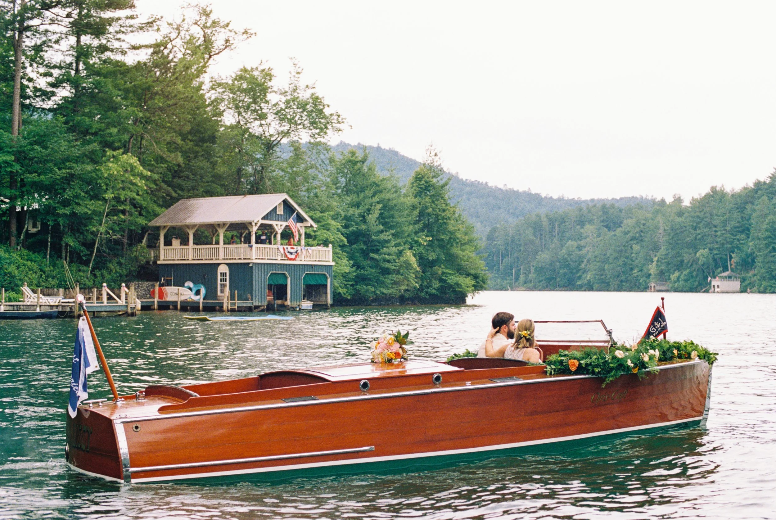 Couple arriving by classic wooden boat at a lakeside boathouse surrounded by trees