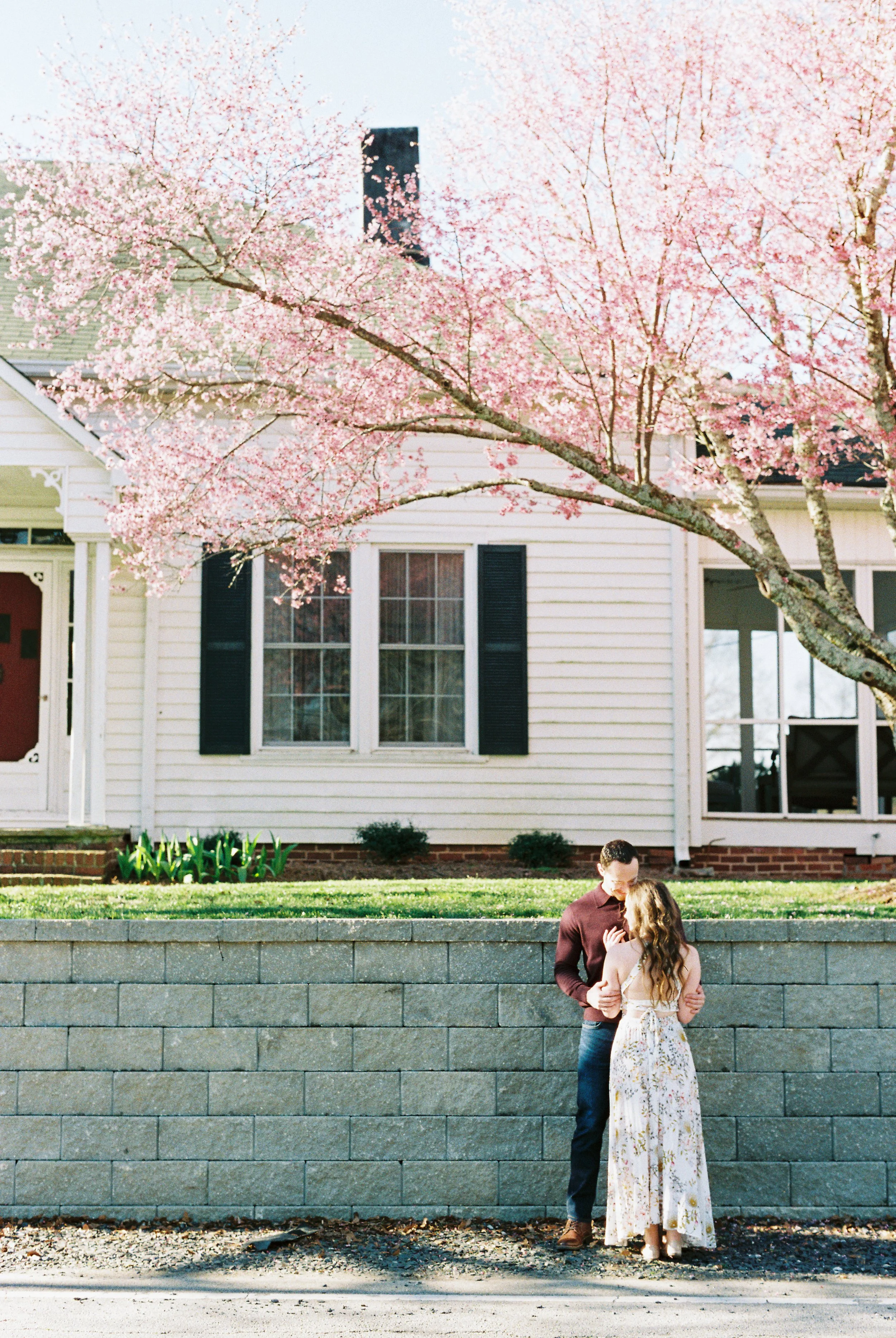Conner and Maddie embrace beside a stone wall beneath a blooming cherry blossom tree in Dallas, Georgia.