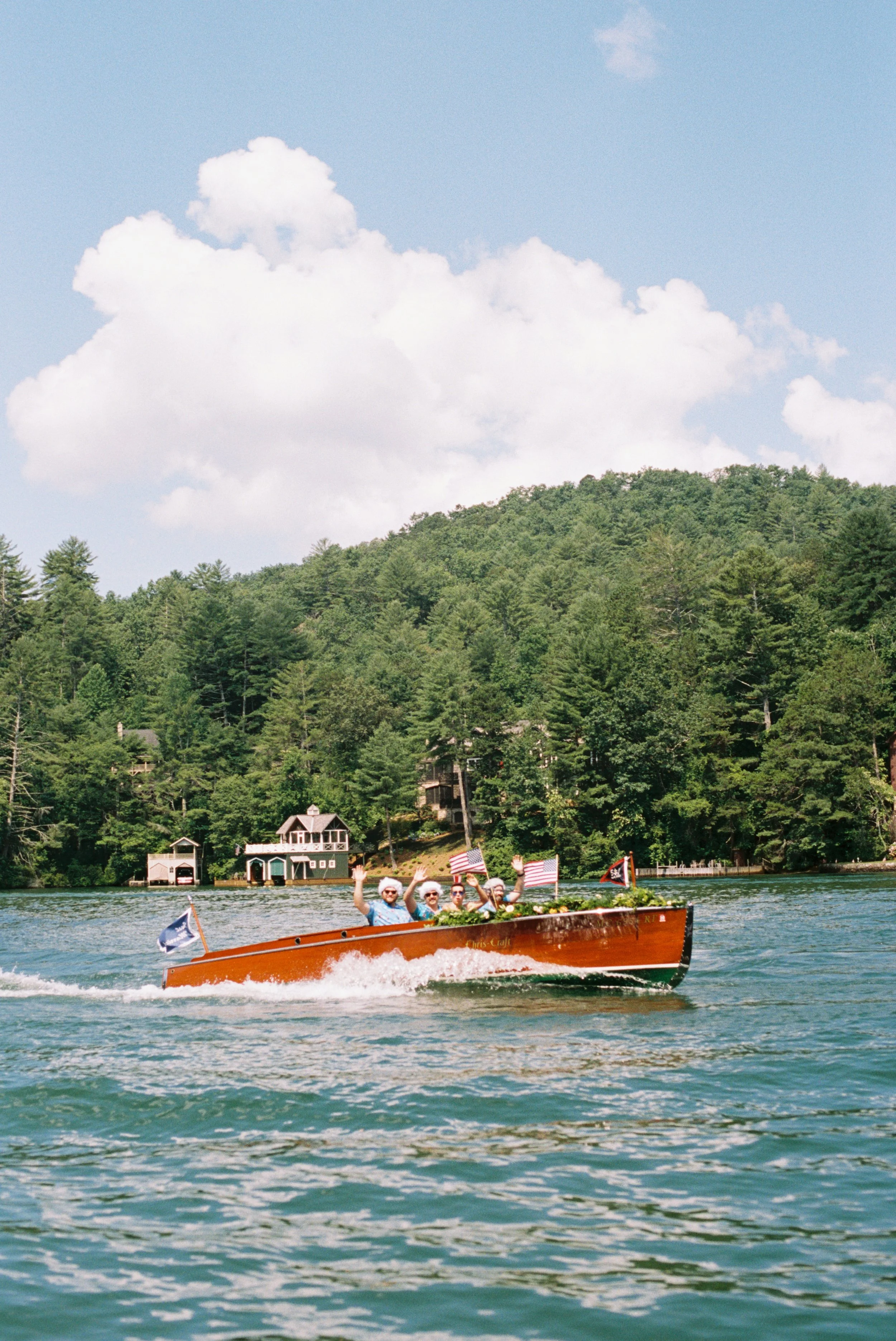 Red wooden boat carrying wedding guests across the lake beneath a blue sky with a boathouse in the distance