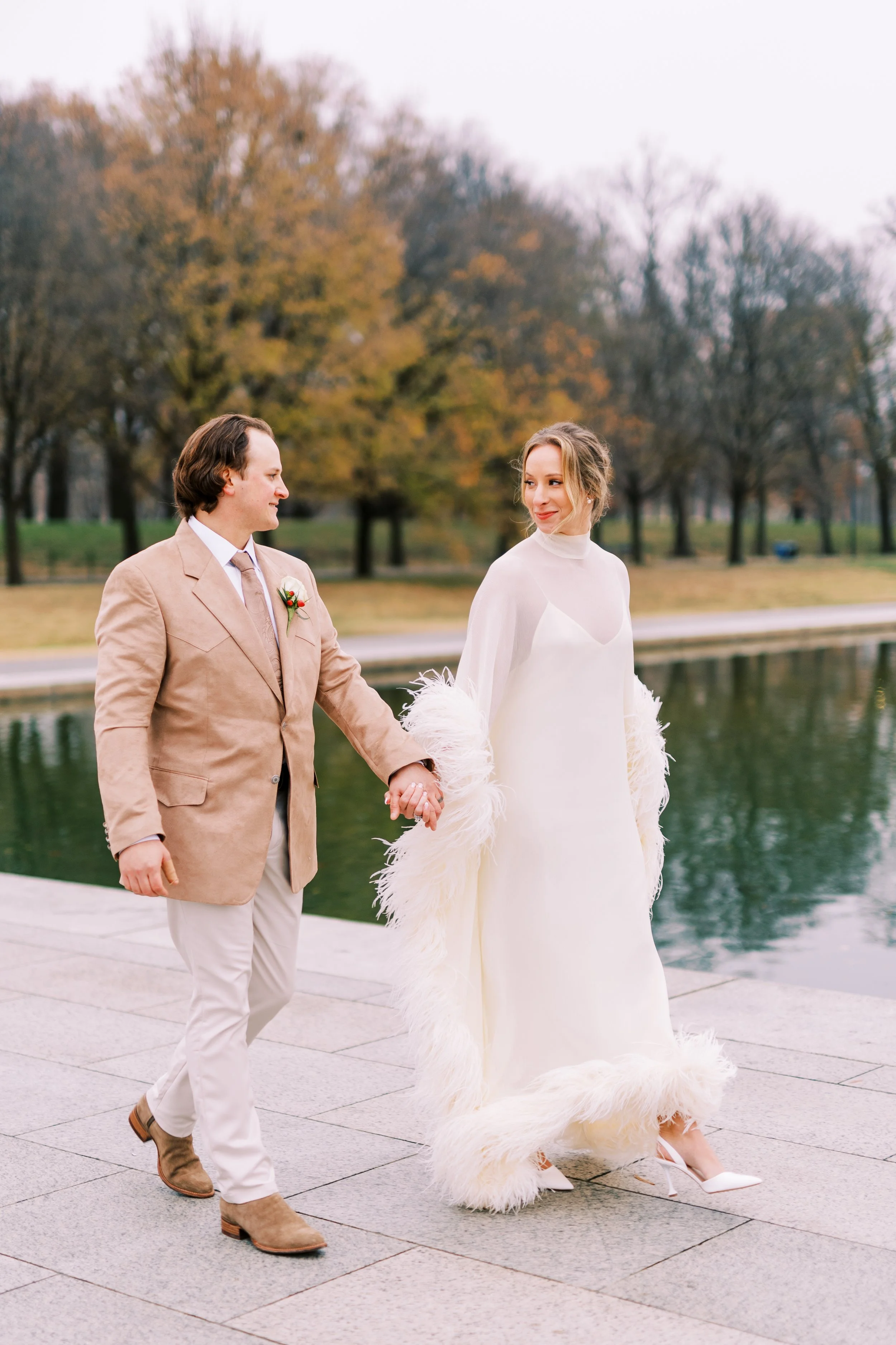 Bride and groom walking hand in hand along the waterfront with autumn trees in the background