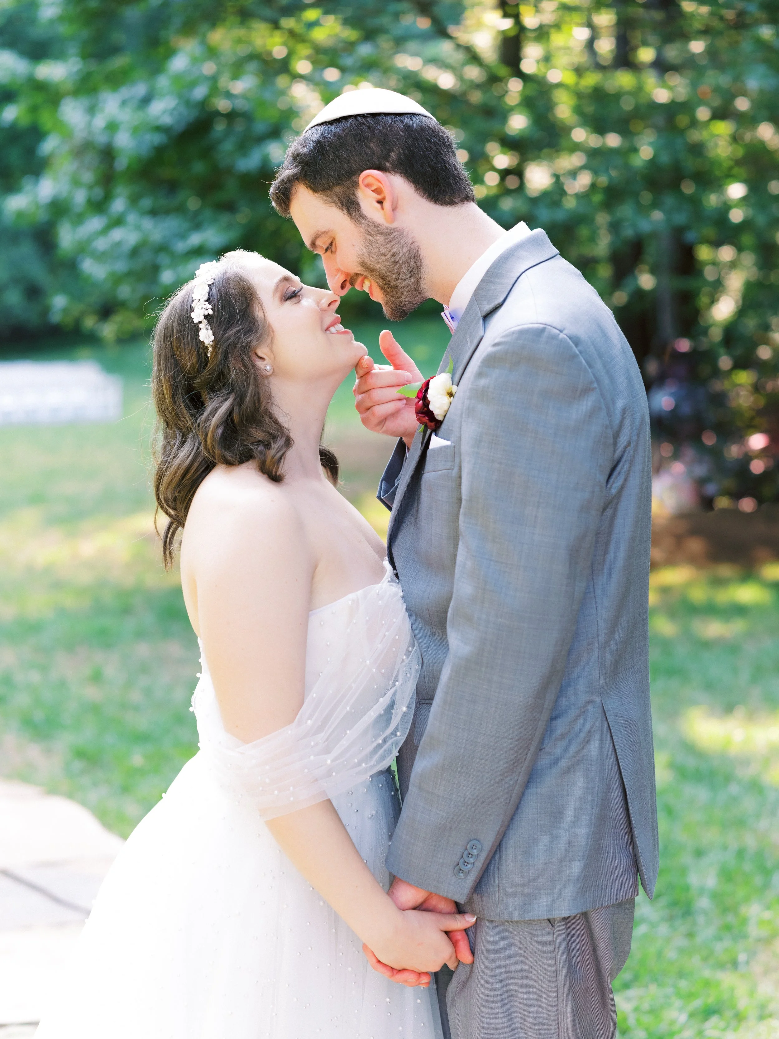 A bride and groom romantically close outdoors during a wedding, holding hands, smiling, with trees and greenery in the background.