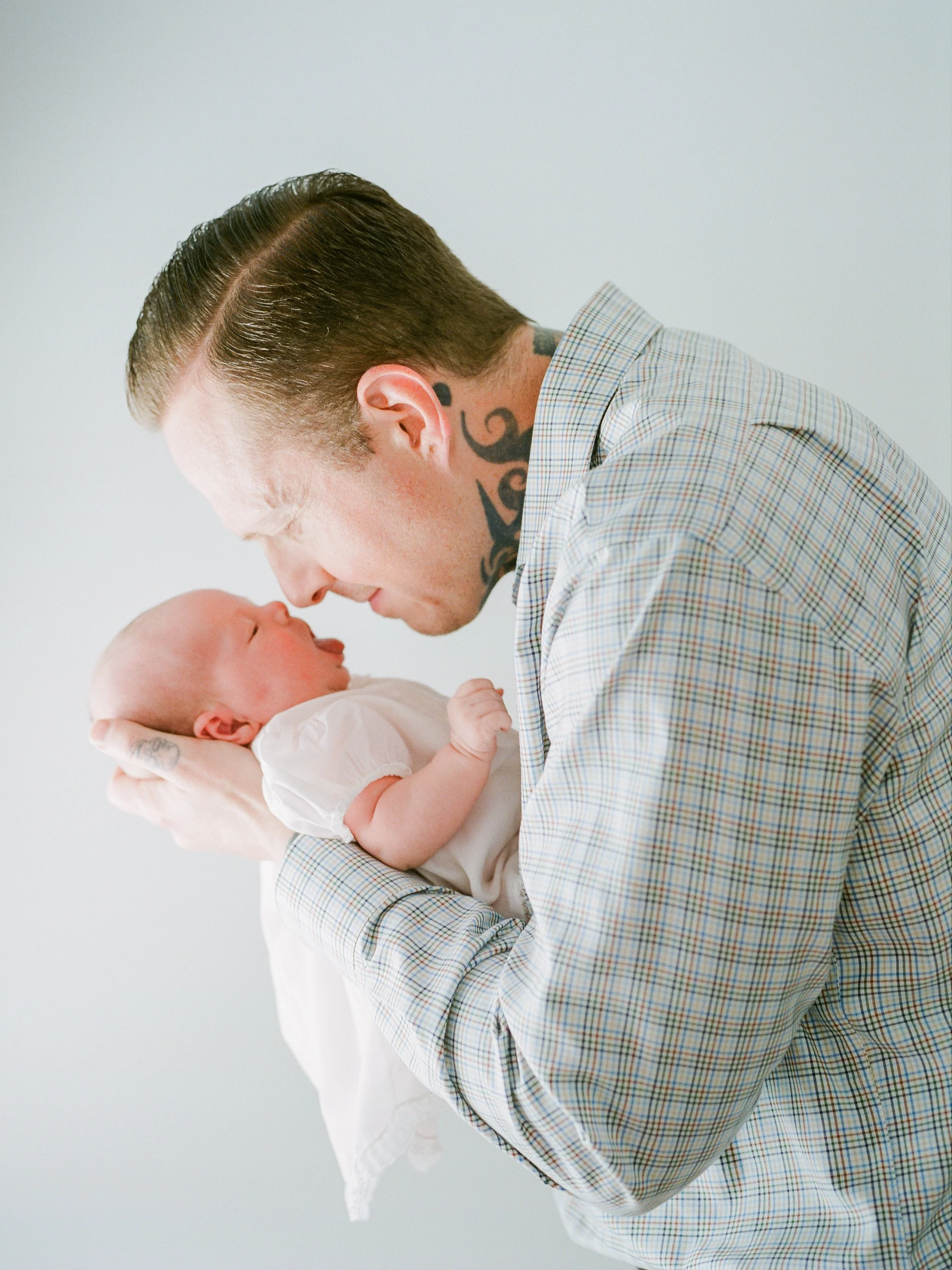 A man with tattoos and a checkered shirt gently holding and touching noses with a newborn baby.