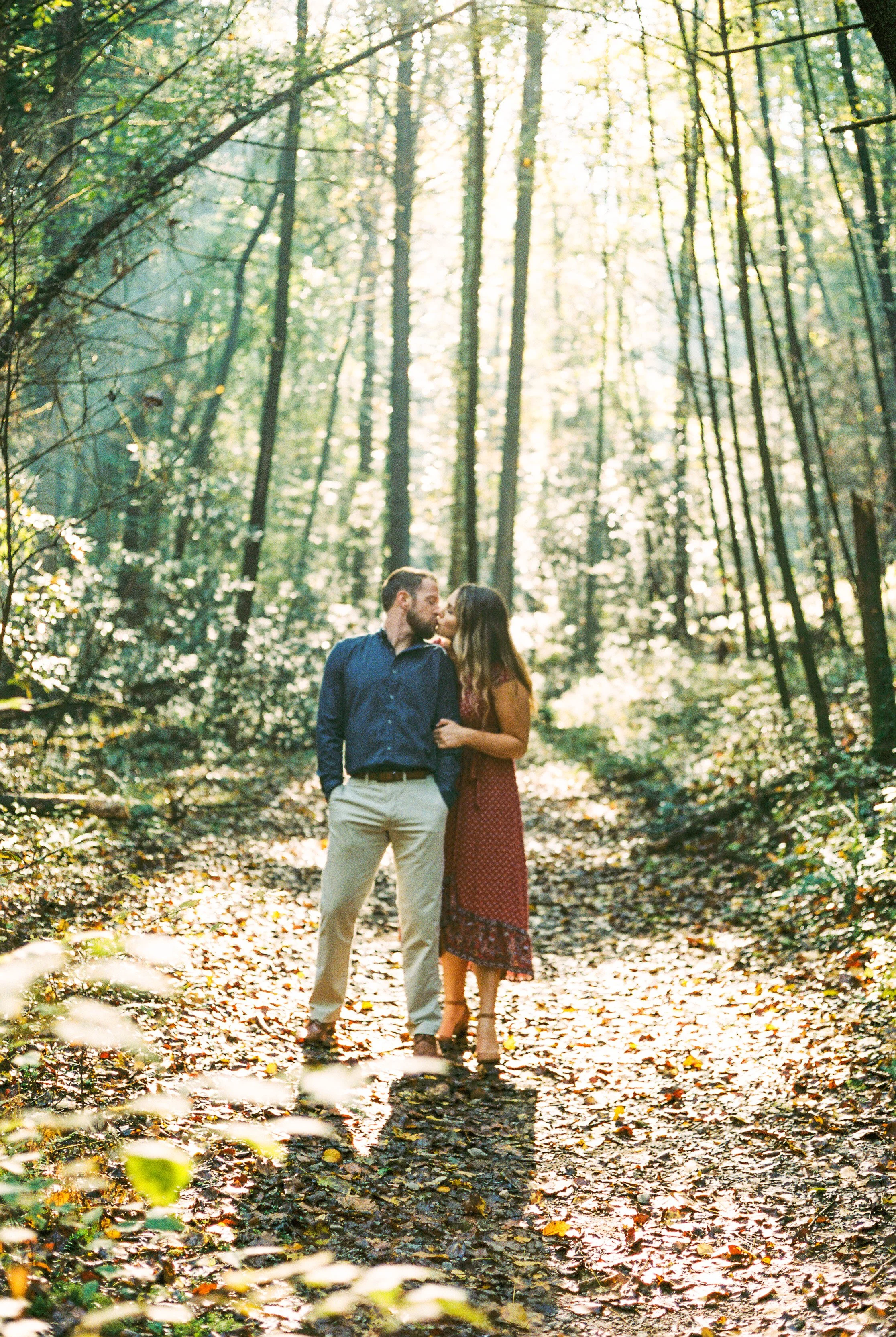 A couple kissing in a sunlit forest with tall trees and fallen leaves on the ground.