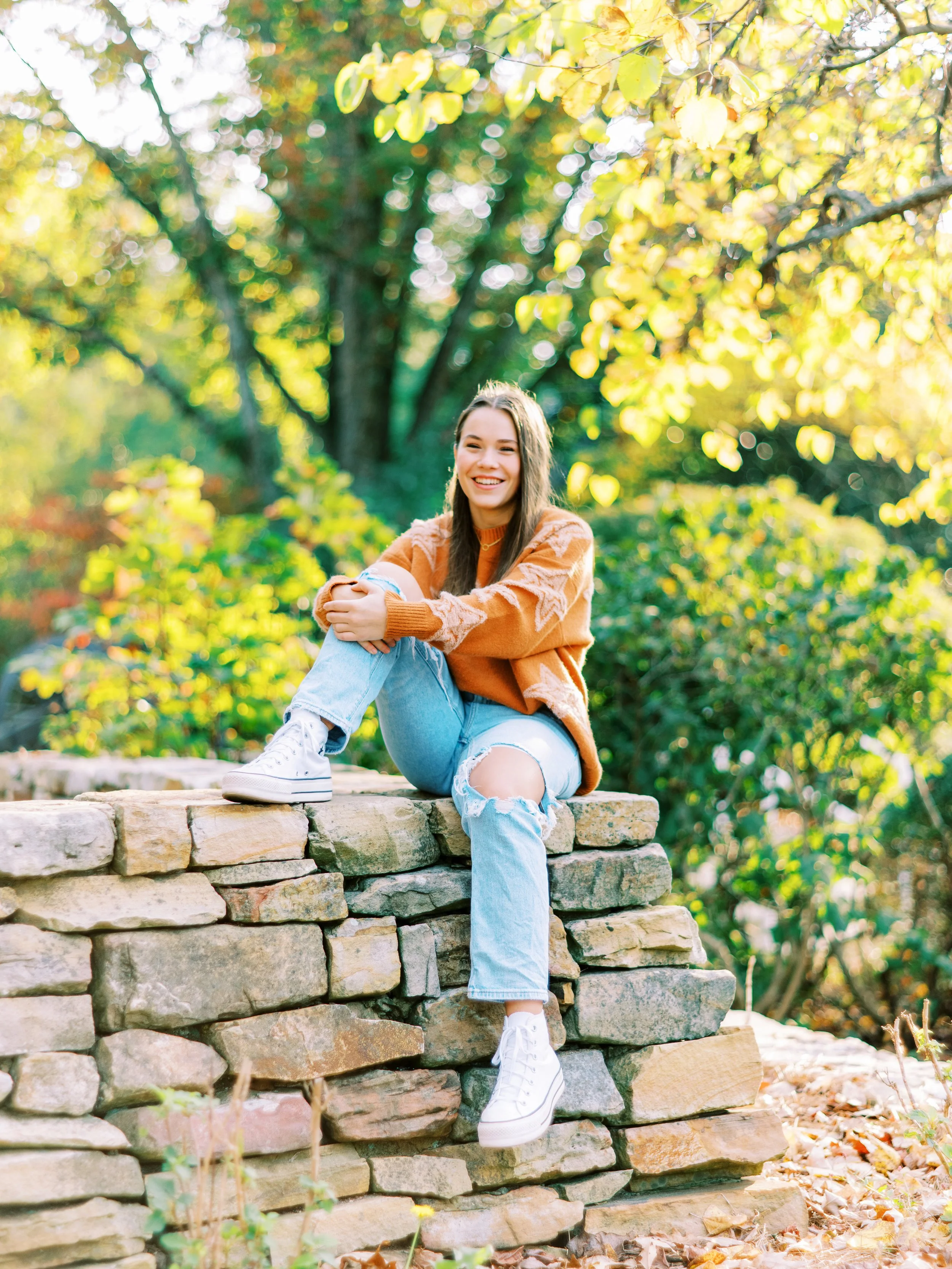 Senior portrait of Hannah sitting on a stone wall with arms crossed over her knees in an orange sweater at Kennesaw Mountain Battlefield Park in Kennesaw, Georgia