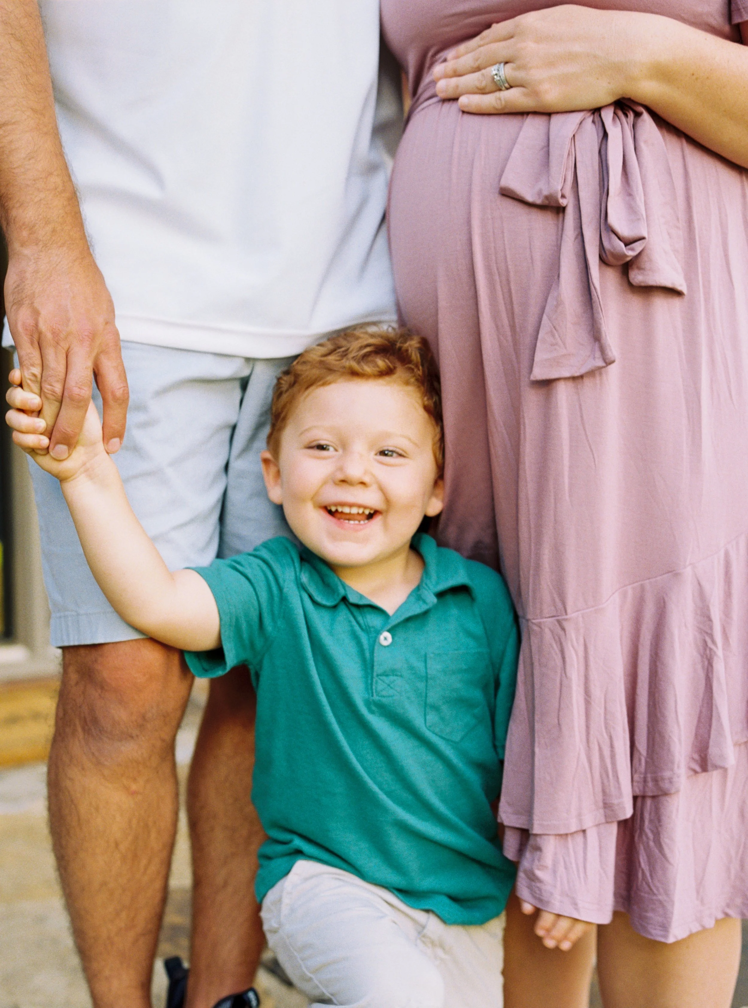 A young boy with red hair and a big smile, wearing a teal shirt and light-colored shorts, is hugging a woman who is wearing a pink dress. They are outdoors, and only parts of the adults are visible, including the man's arm on the left holding the boy's hand.