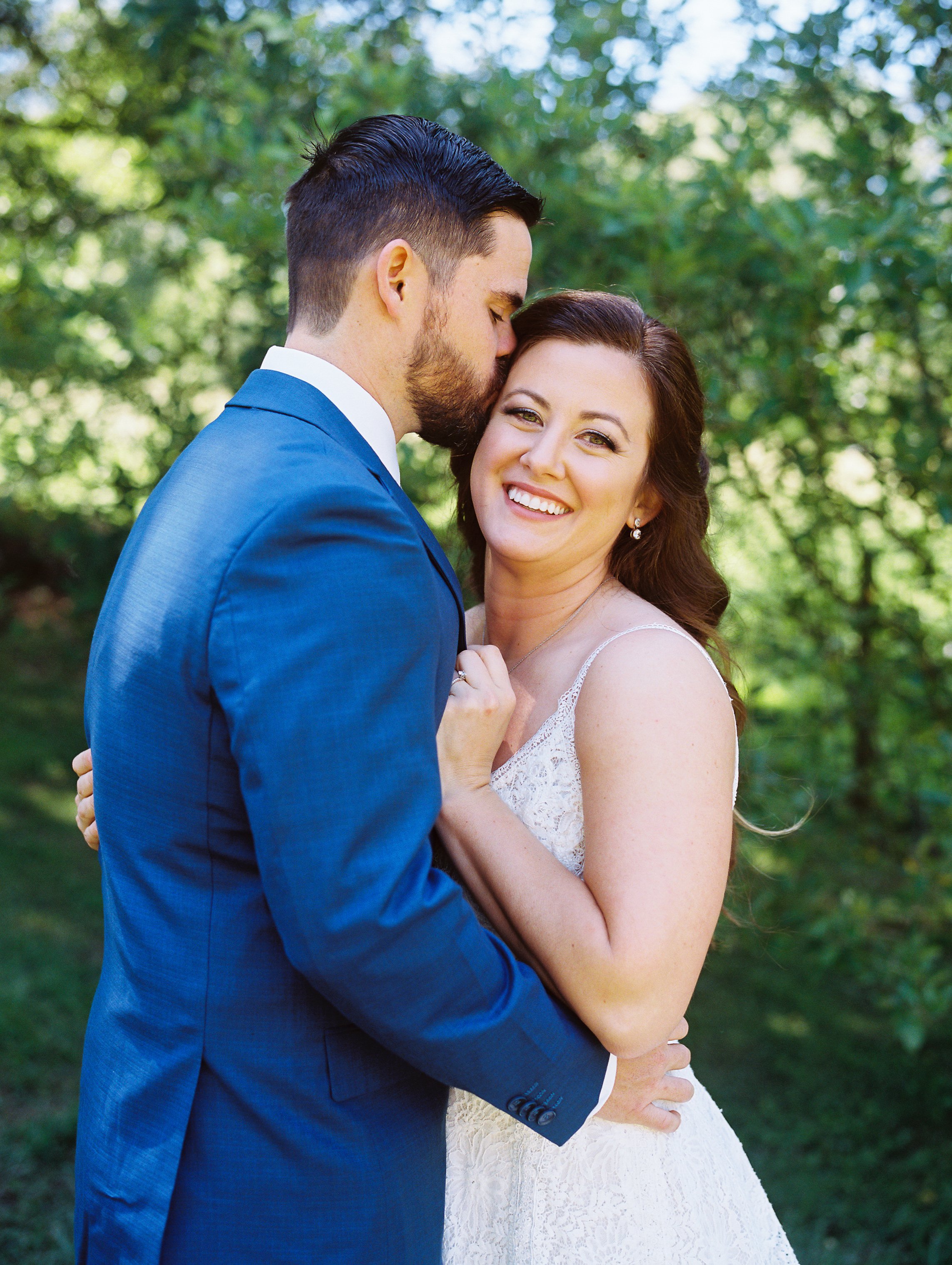 Groom kissing the bride's cheek while embracing her outdoors, bride smiling at the camera