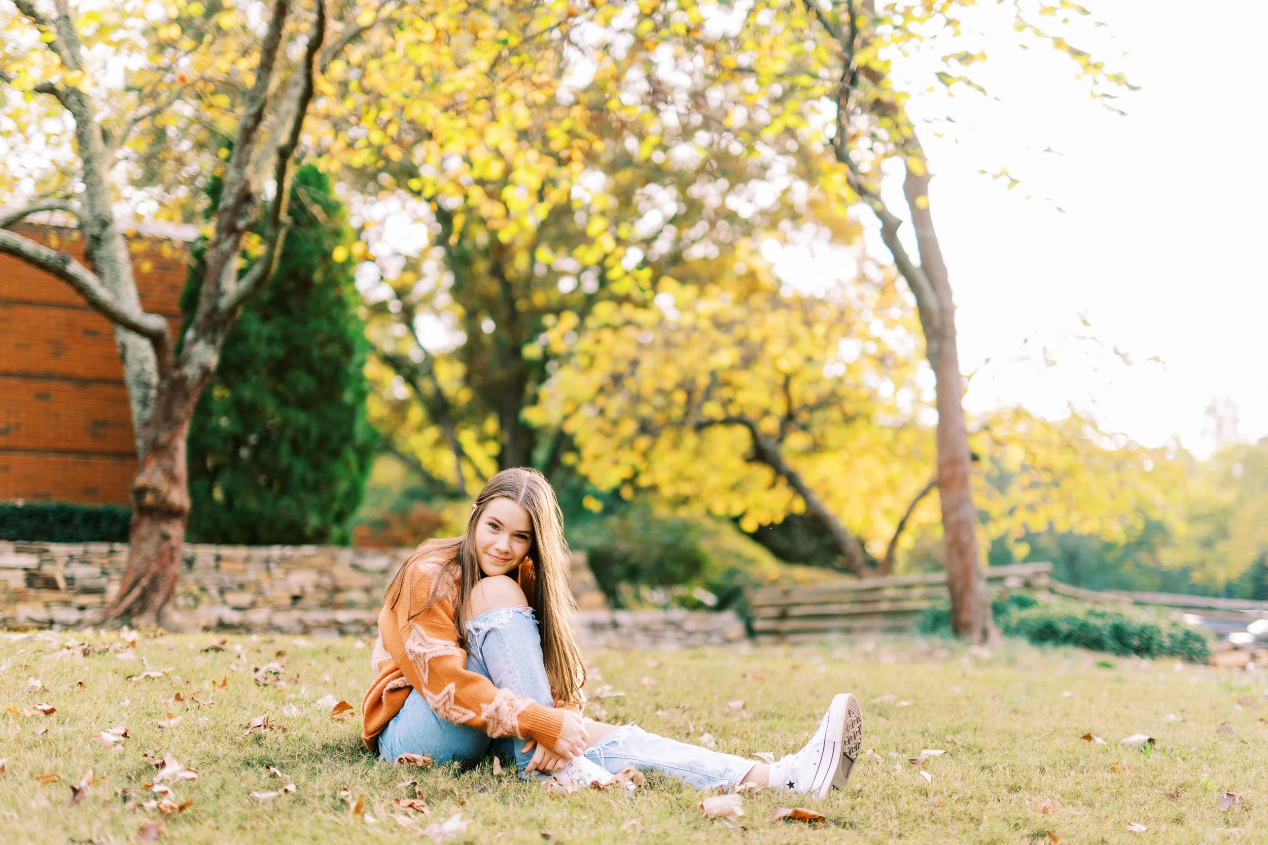 Wide senior portrait of Hannah posing on the grass near a red barn in an orange sweater at Kennesaw Mountain Battlefield Park in Kennesaw, Georgia