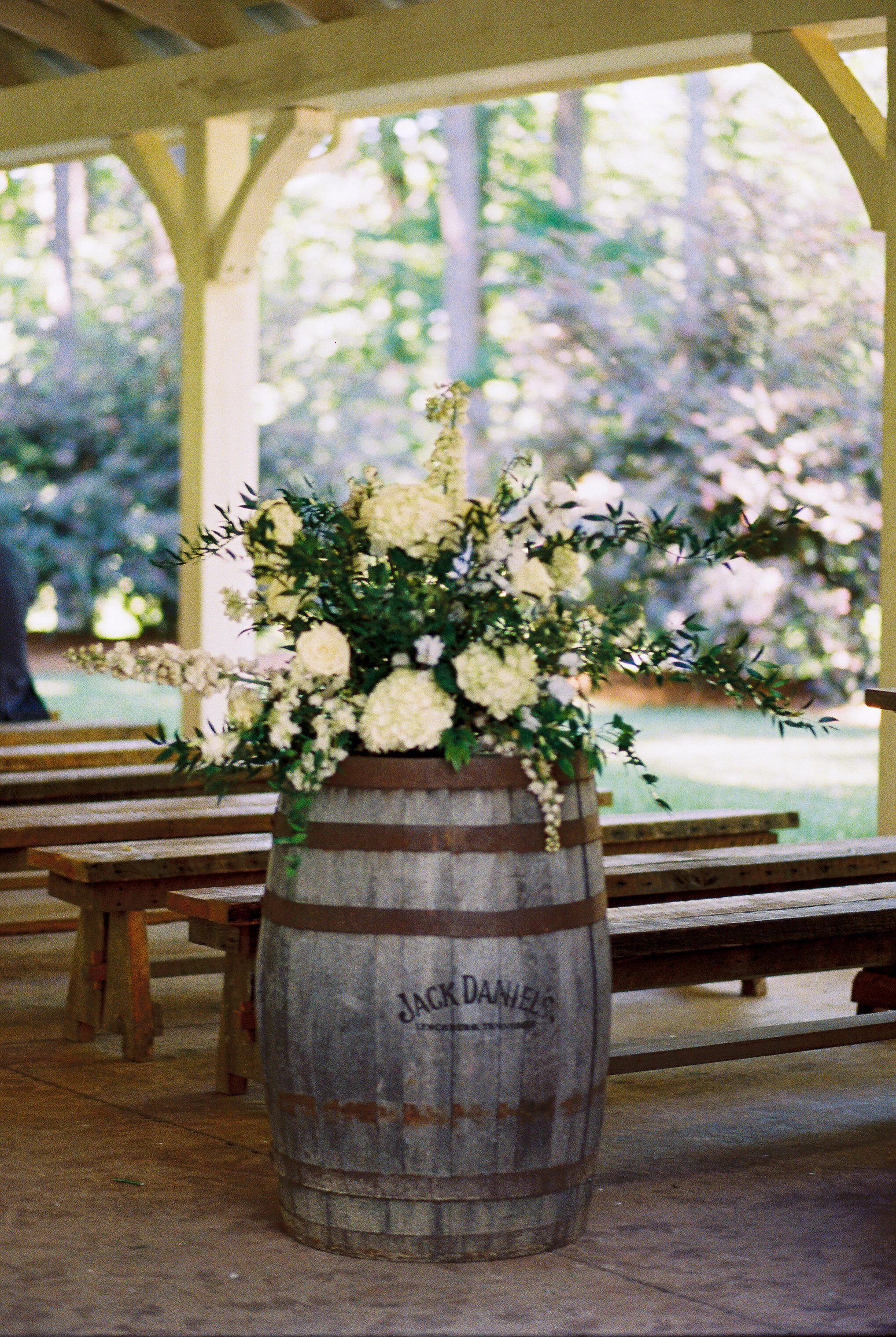 Floral centerpiece of white and cream flowers displayed atop a wooden whiskey barrel near the wedding venue's wooden benches