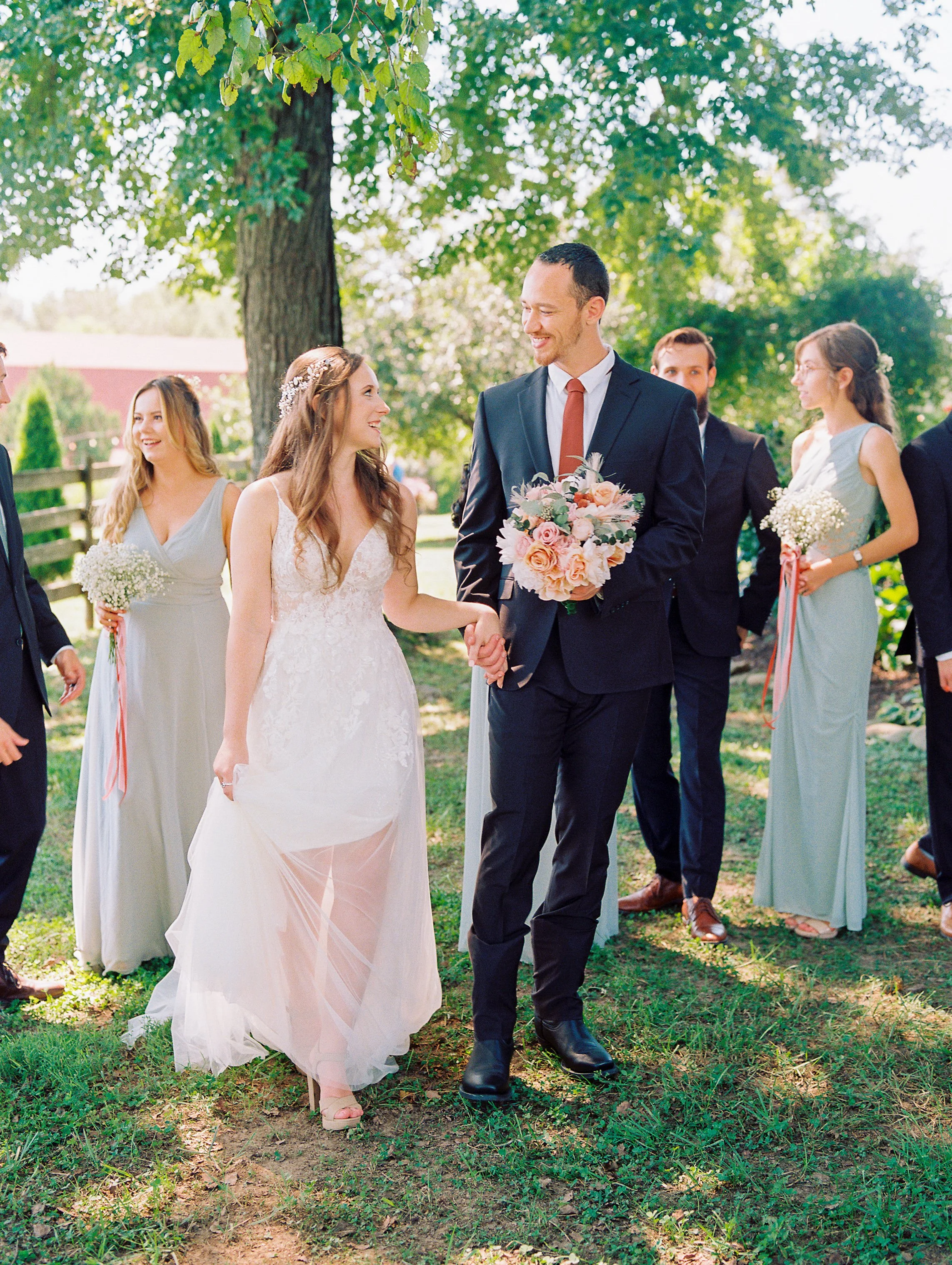 Wedding ceremony outdoors with bride and groom holding hands, surrounded by bridesmaids and groomsmen under trees.
