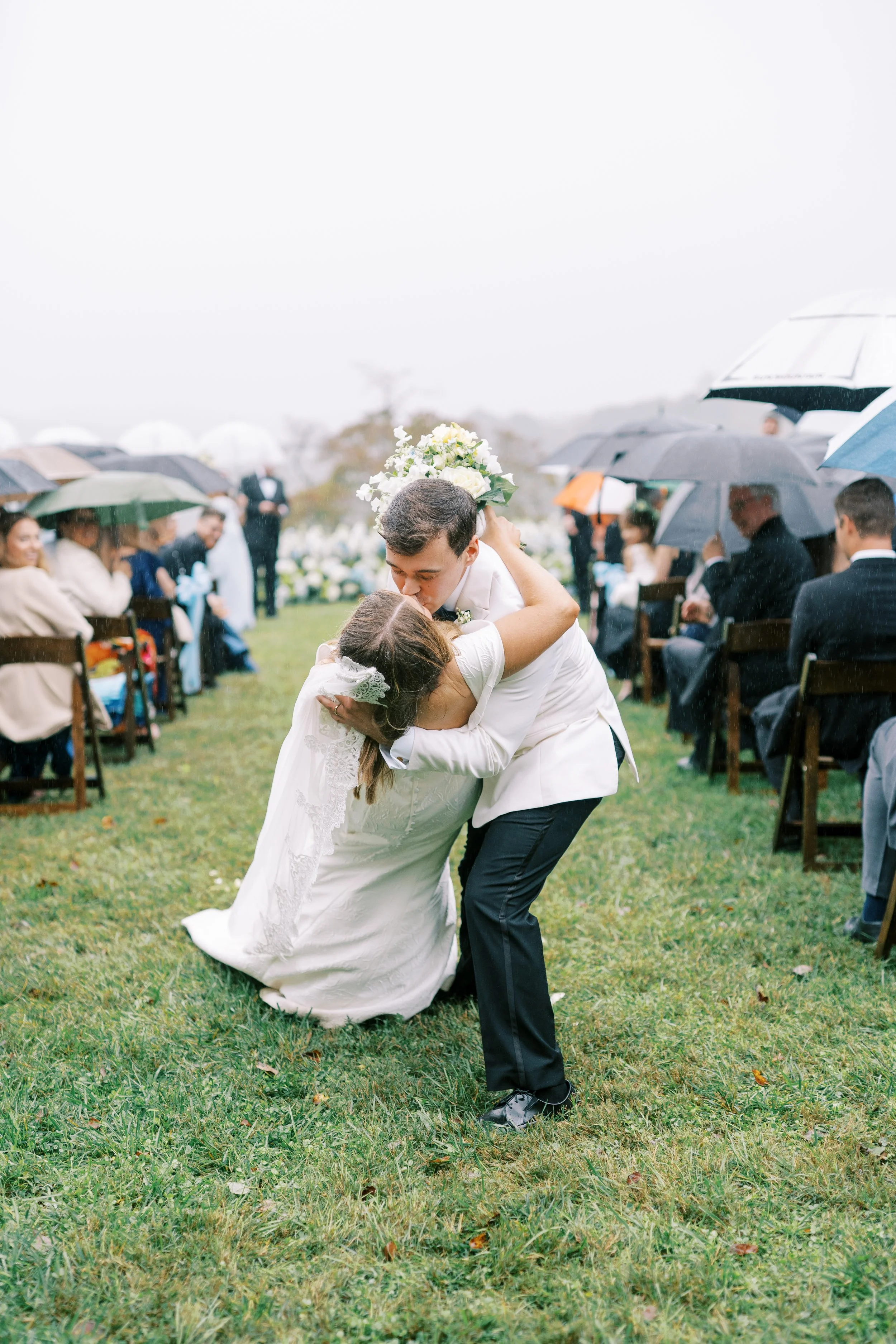 Groom playfully dipping bride during reception with umbrella-holding guests in the background