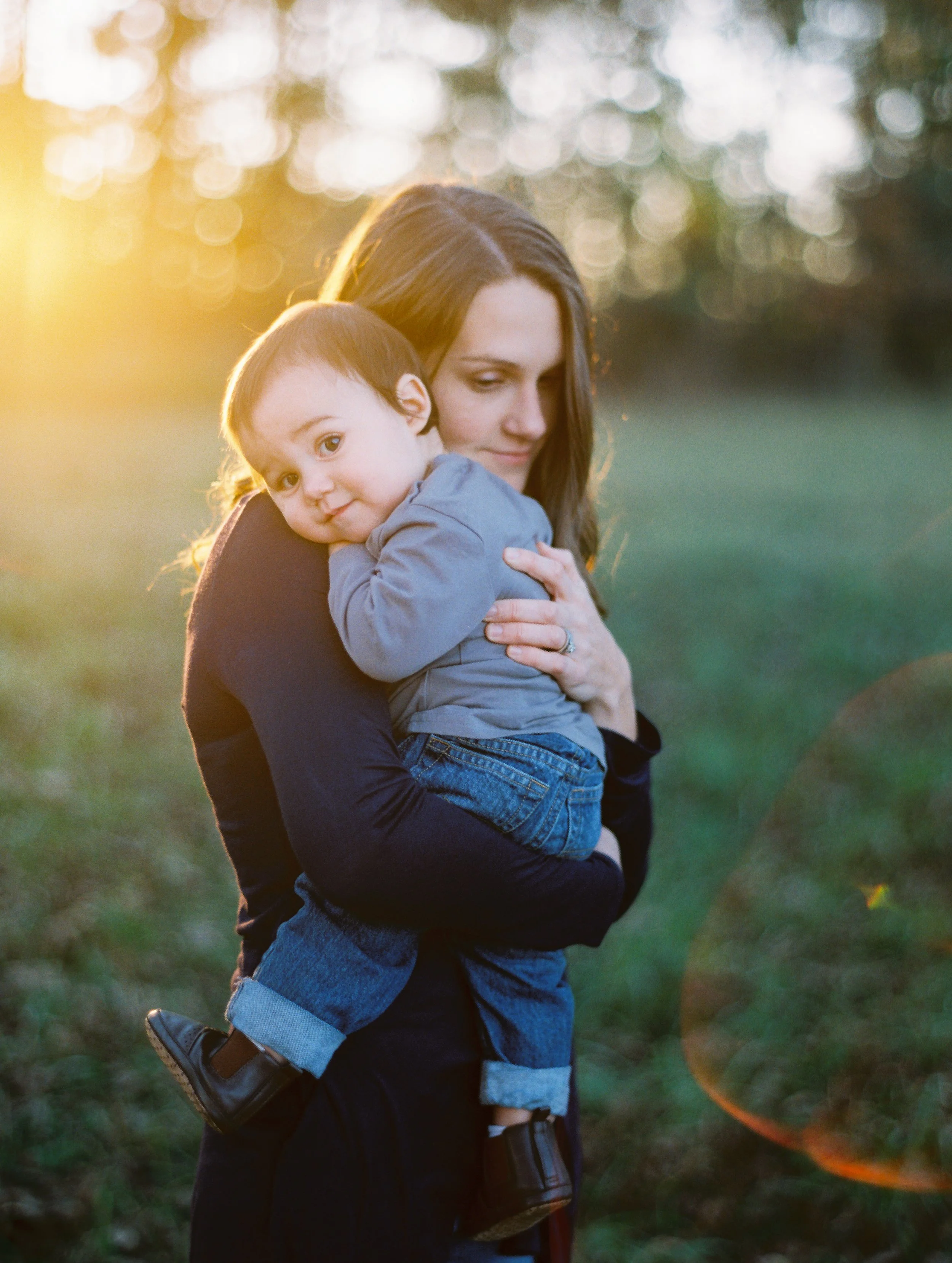 A woman holding a young child in an outdoor setting during sunset, with trees in the background and a lens flare at her mommy and me portrait session with her toddler son.