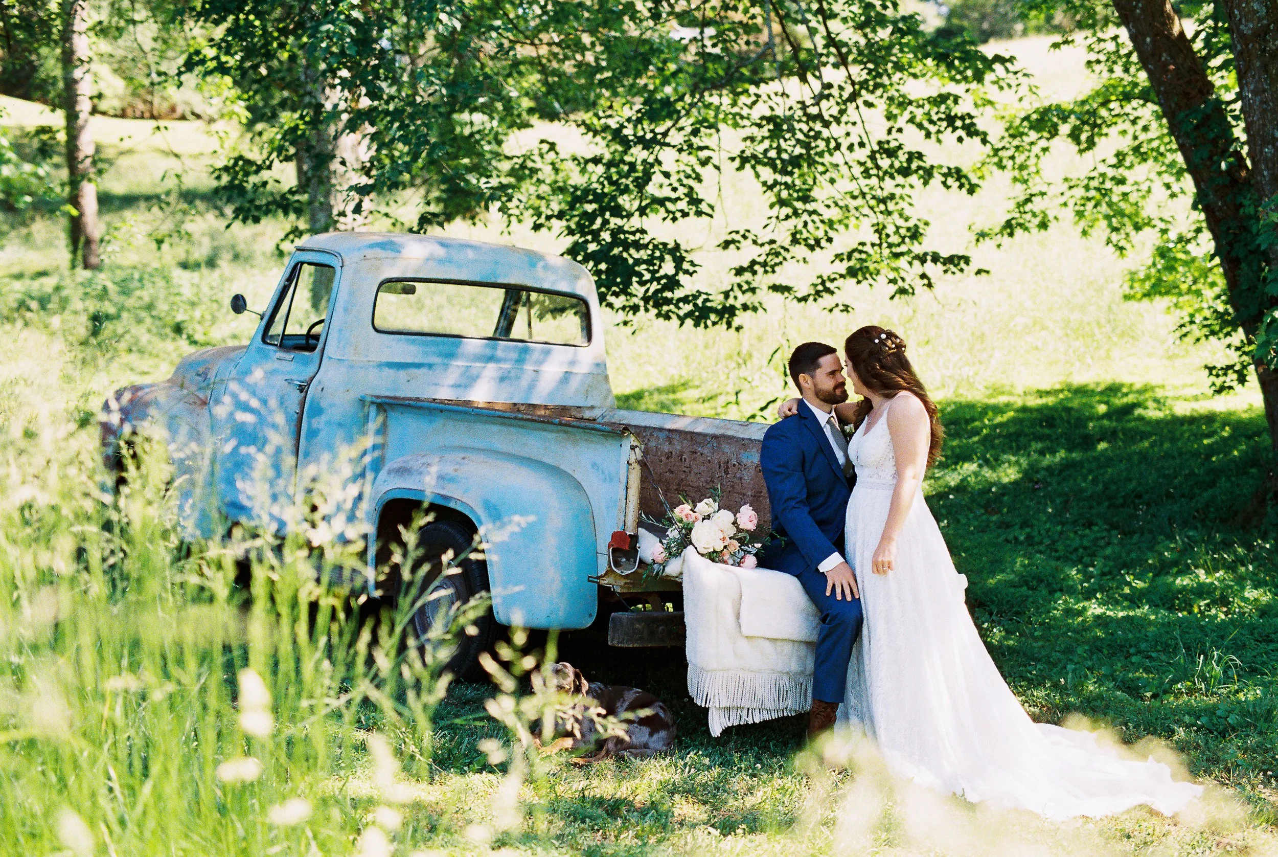 Bride and groom embracing beside the vintage blue truck, shot through tall grass in the foreground, bouquet resting on the tailgate