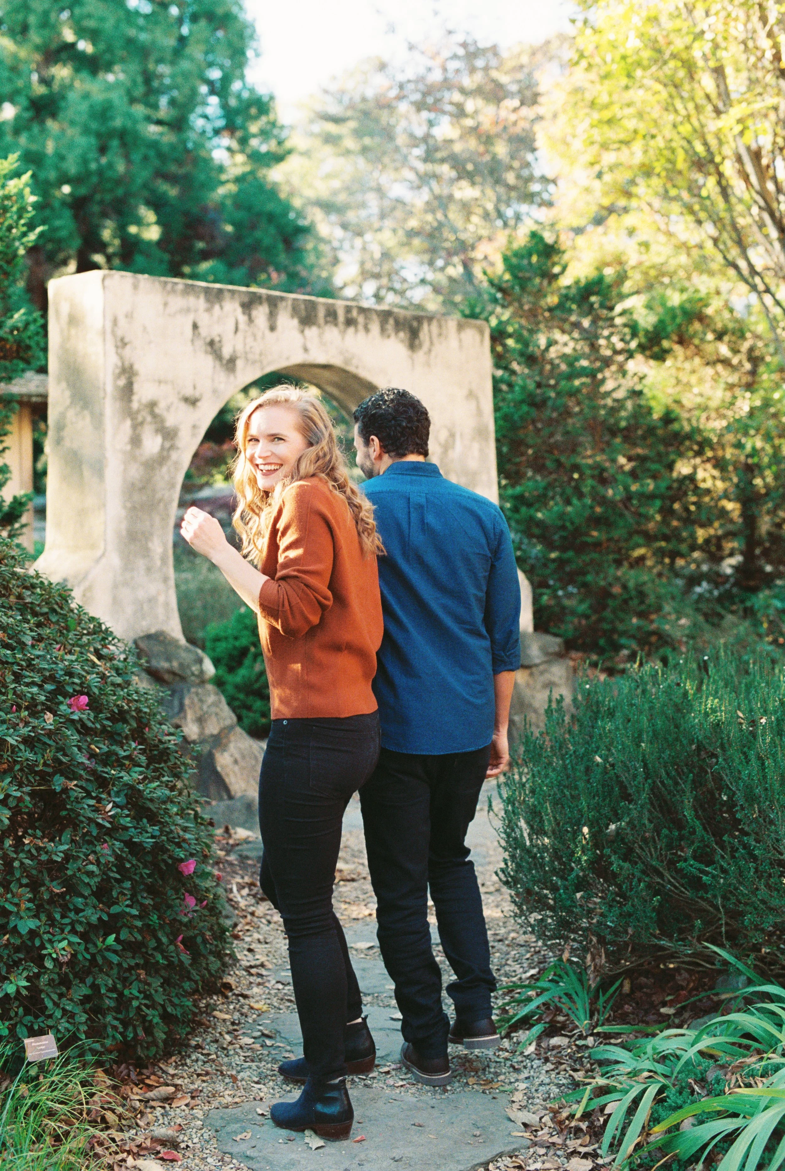 A woman with long, wavy hair smiling and looking back, while a man walks behind her in a lush garden with green shrubs and trees, with a stone archway in the background at Atlanta Botanical Gardens.