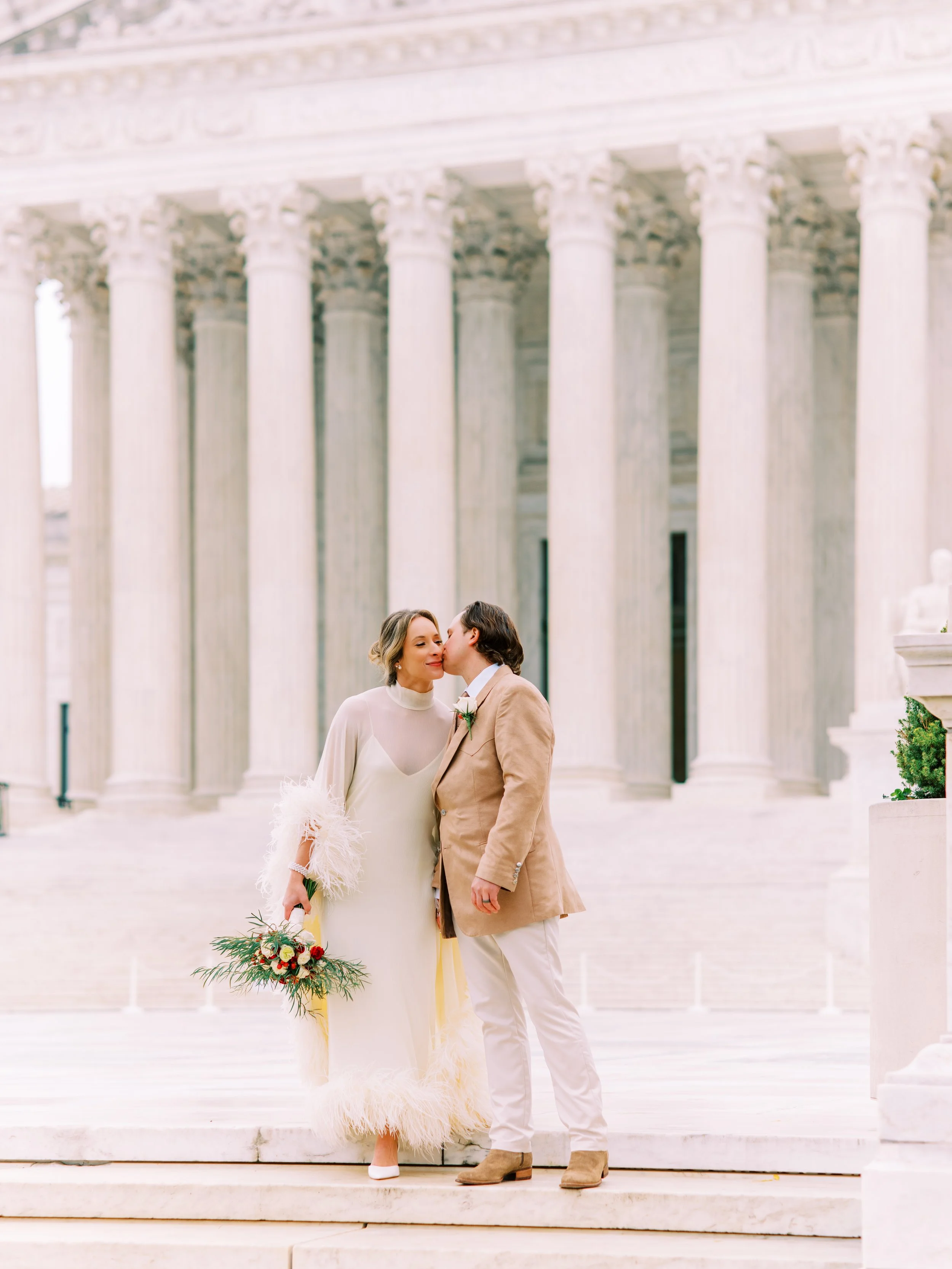 Bride and groom posing together in front of a grand columned building, bride holding her bouquet