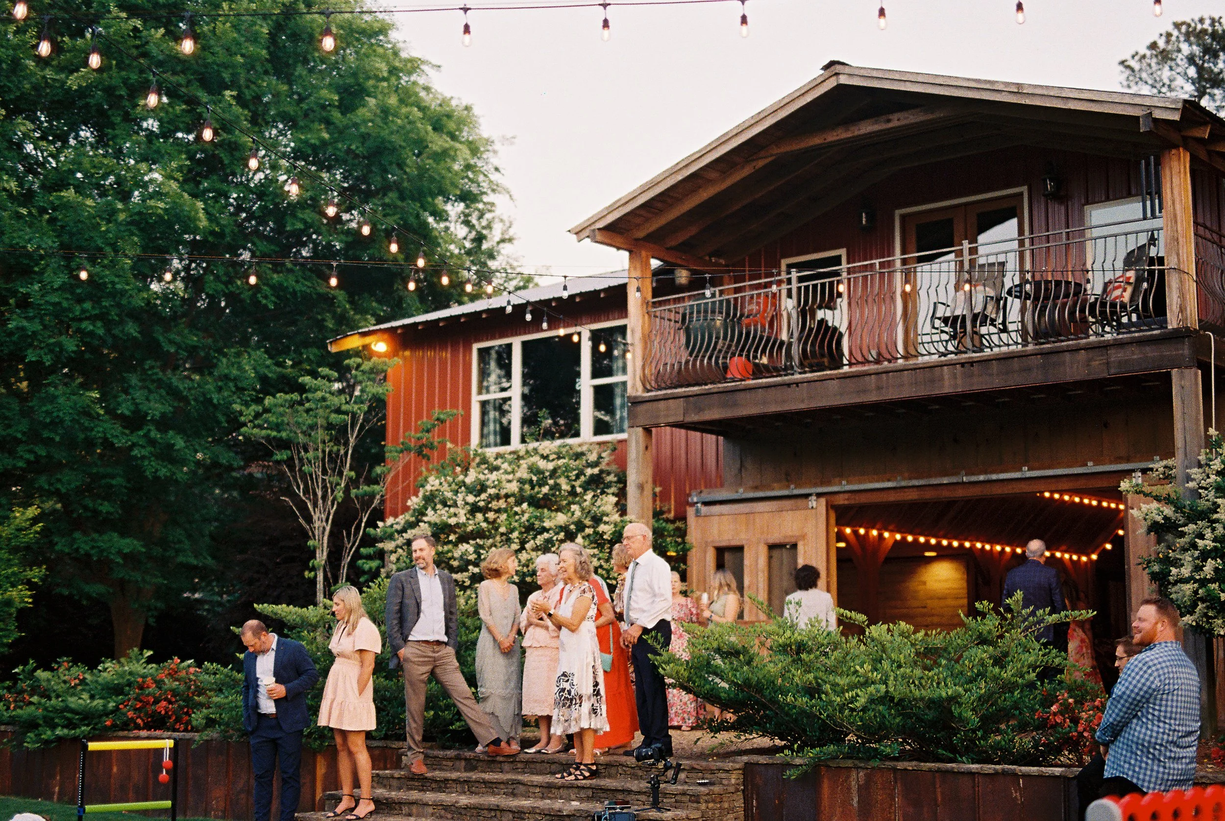 Guests mingling on the patio outside the illuminated red barn venue at dusk, string lights strung along the balcony and eaves