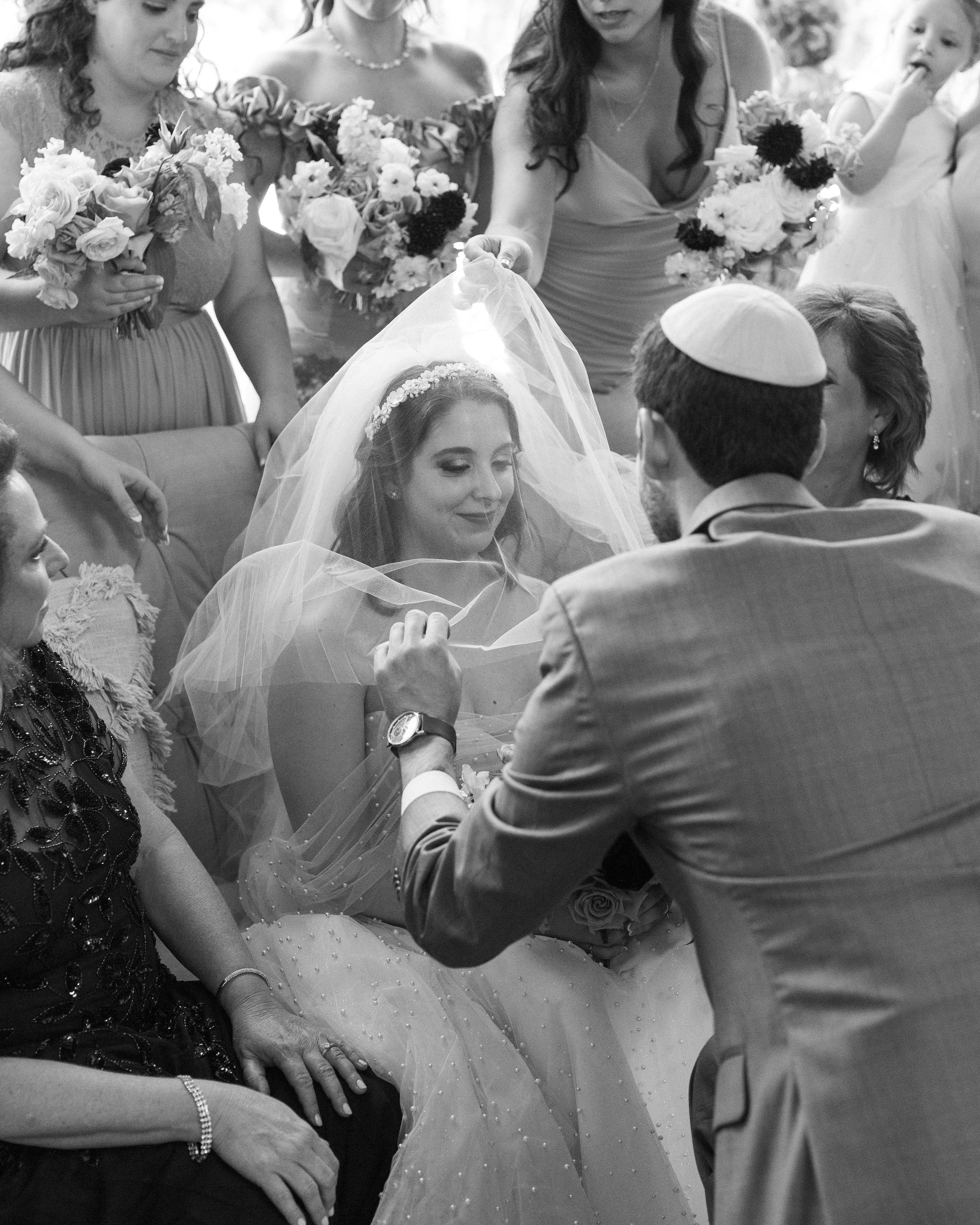 A bride in a wedding dress with a veil and floral headpiece, sitting and smiling as a man in a yarmulke adjusts her veil during her wedding ceremony, surrounded by women holding bouquets.