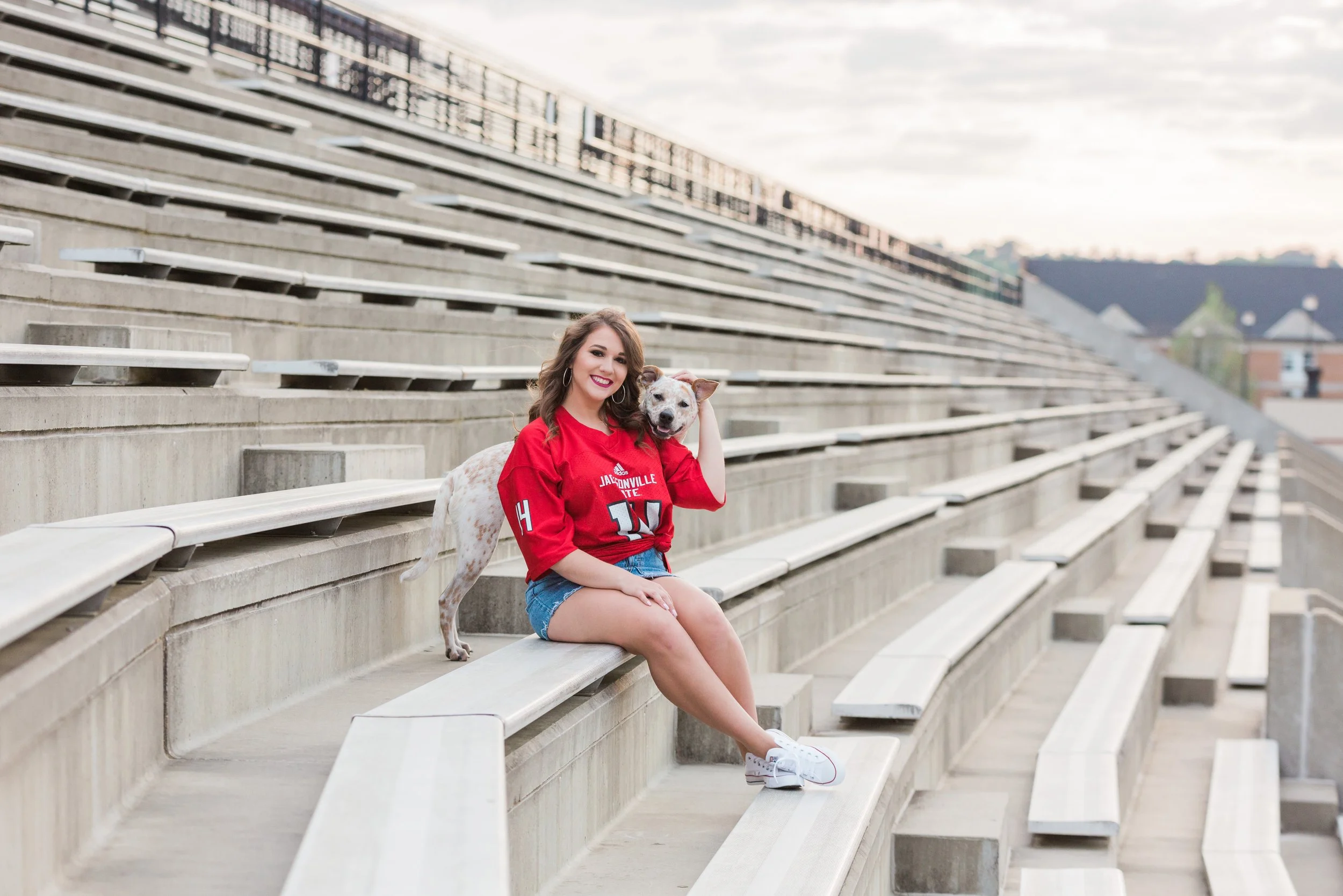 A young woman in a red sports jersey and denim shorts sitting on stadium bleachers with her dog.