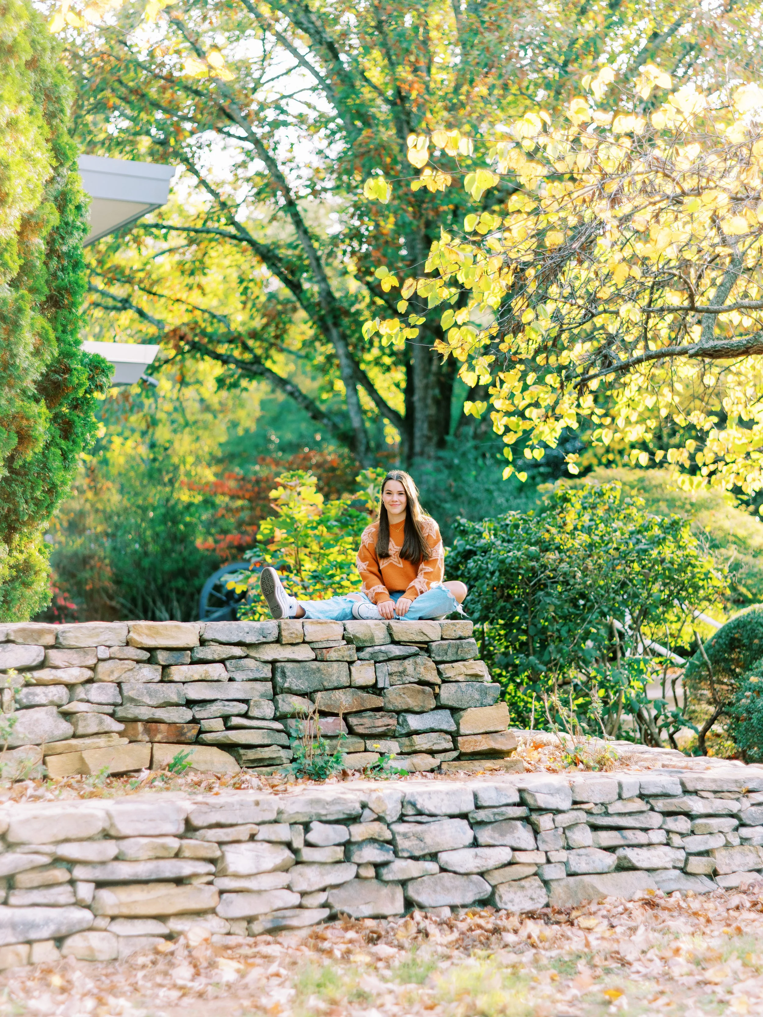 Wide senior portrait of Hannah sitting atop a tiered stone wall in an orange sweater at Kennesaw Mountain Battlefield Park in Kennesaw, Georgia