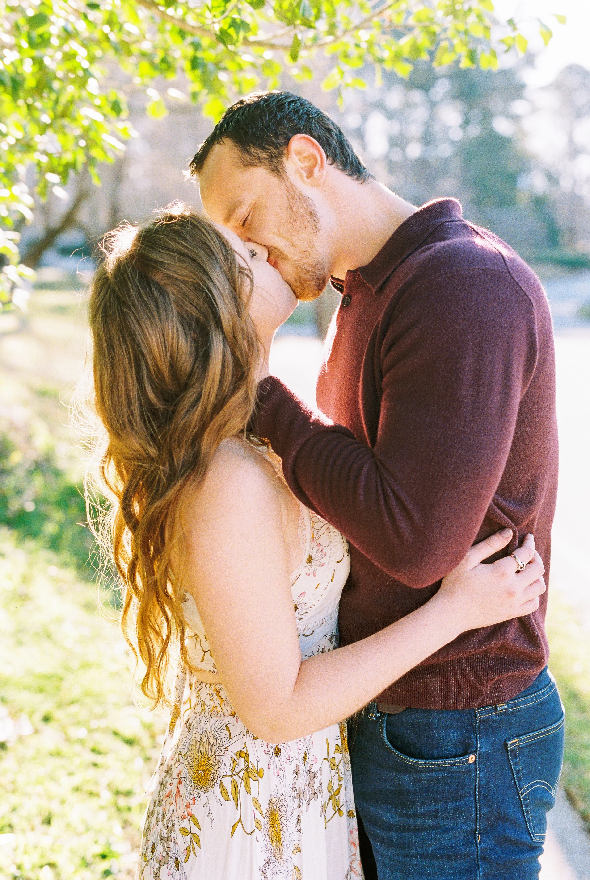 Conner and Maddie kiss beneath dappled tree light beside a path in Dallas, Georgia.