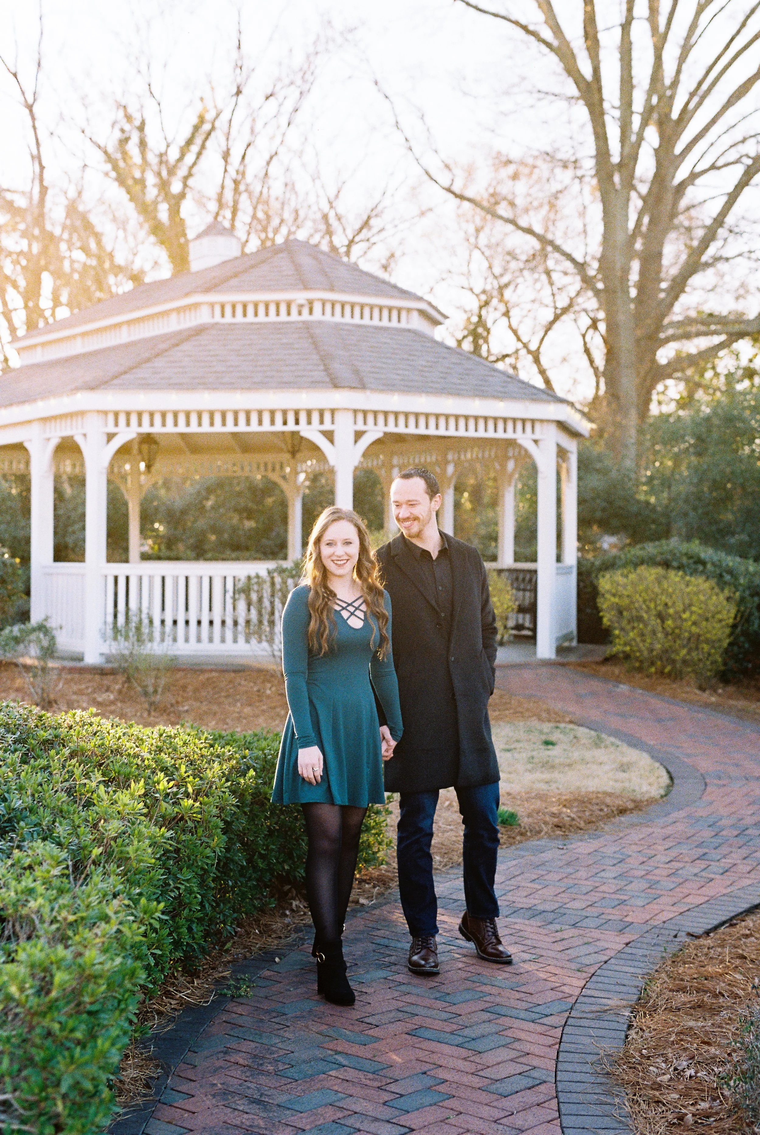 Conner and Maddie walk hand in hand along a brick path near a park gazebo at golden hour in Dallas, Georgia.
