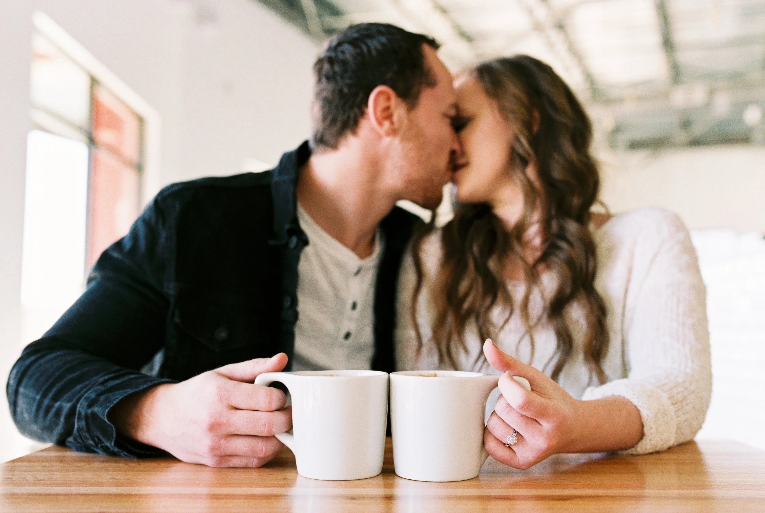 Conner and Maddie share a kiss at their coffee table with the shop's industrial ceiling visible above in Dallas, Georgia.