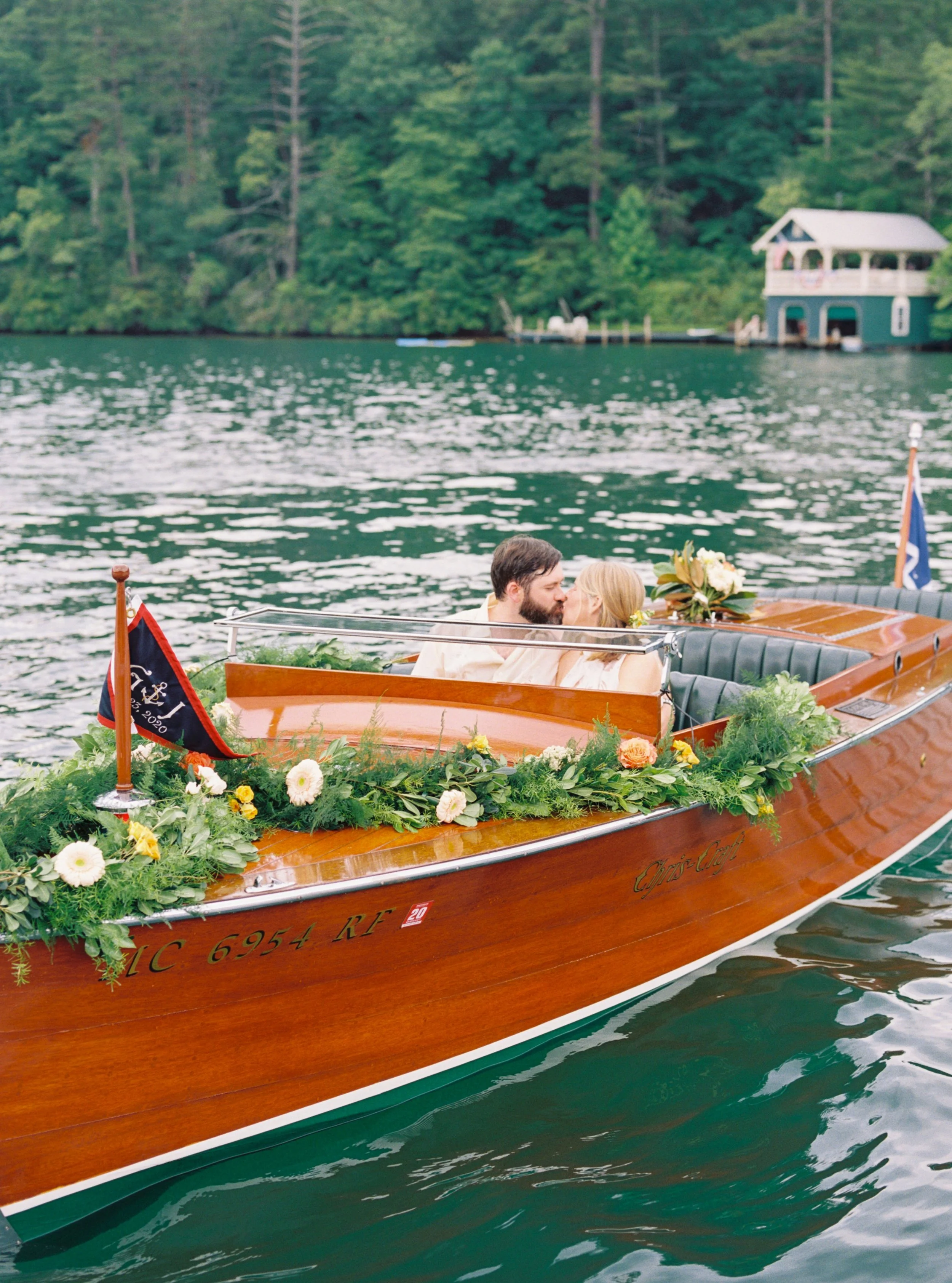 A couple kissing on a decorated wooden boat on a lake, surrounded by greenery and trees.