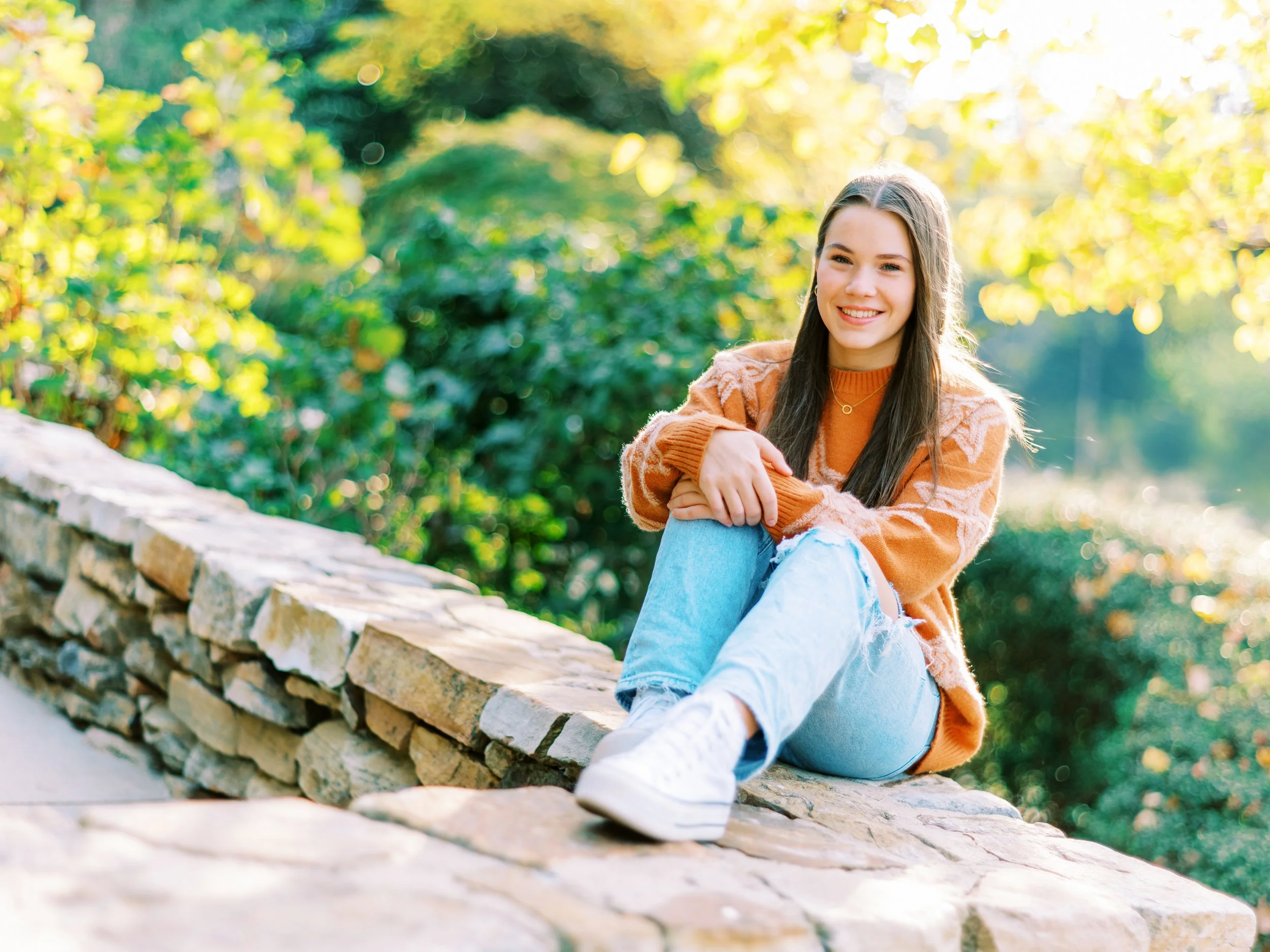 Senior portrait of Hannah smiling while sitting on a stone wall in an orange sweater at Kennesaw Mountain Battlefield Park in Kennesaw, Georgia