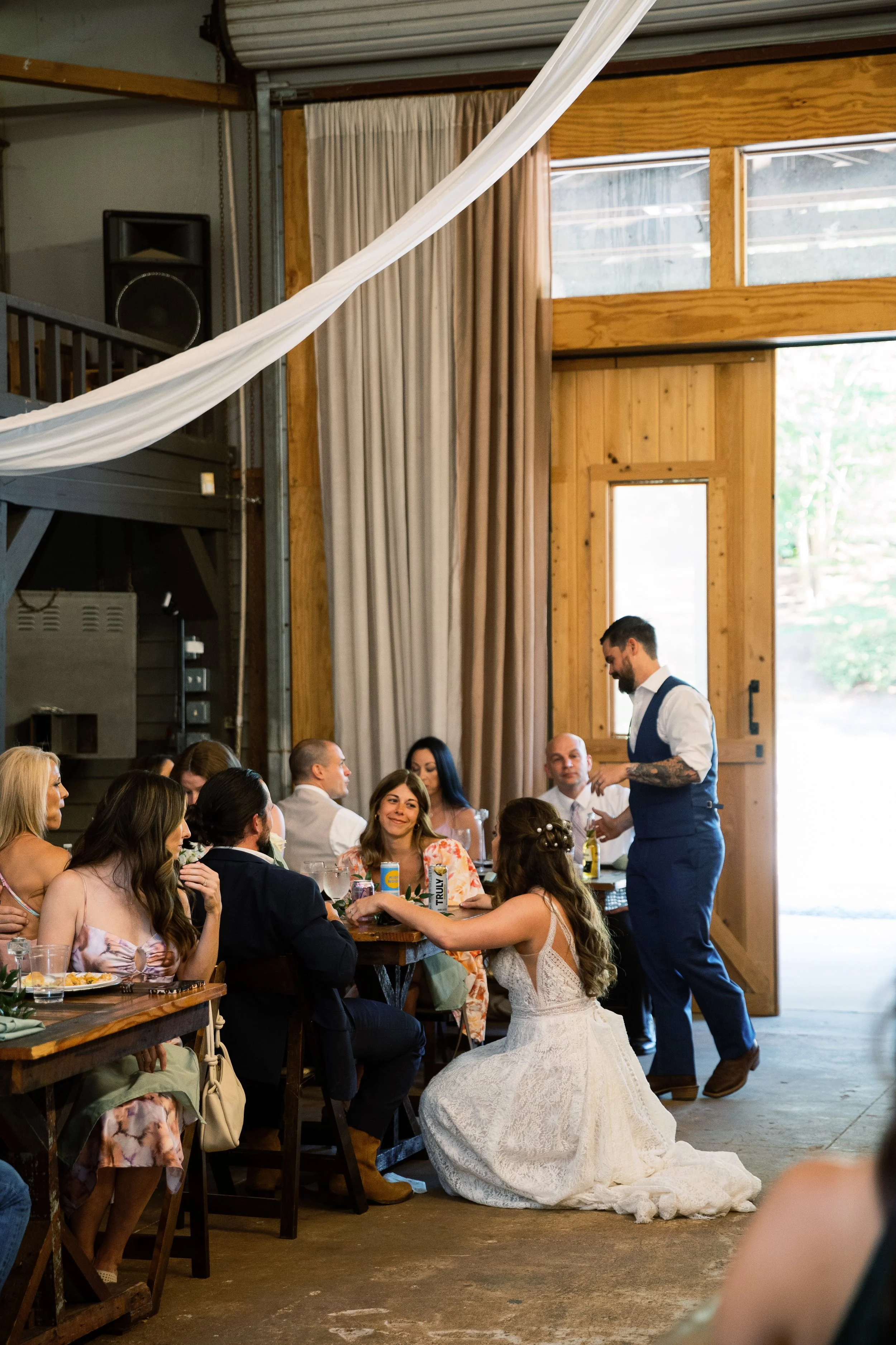 Bride kneeling to greet seated guests at the long reception table indoors as the groom walks by with a drink