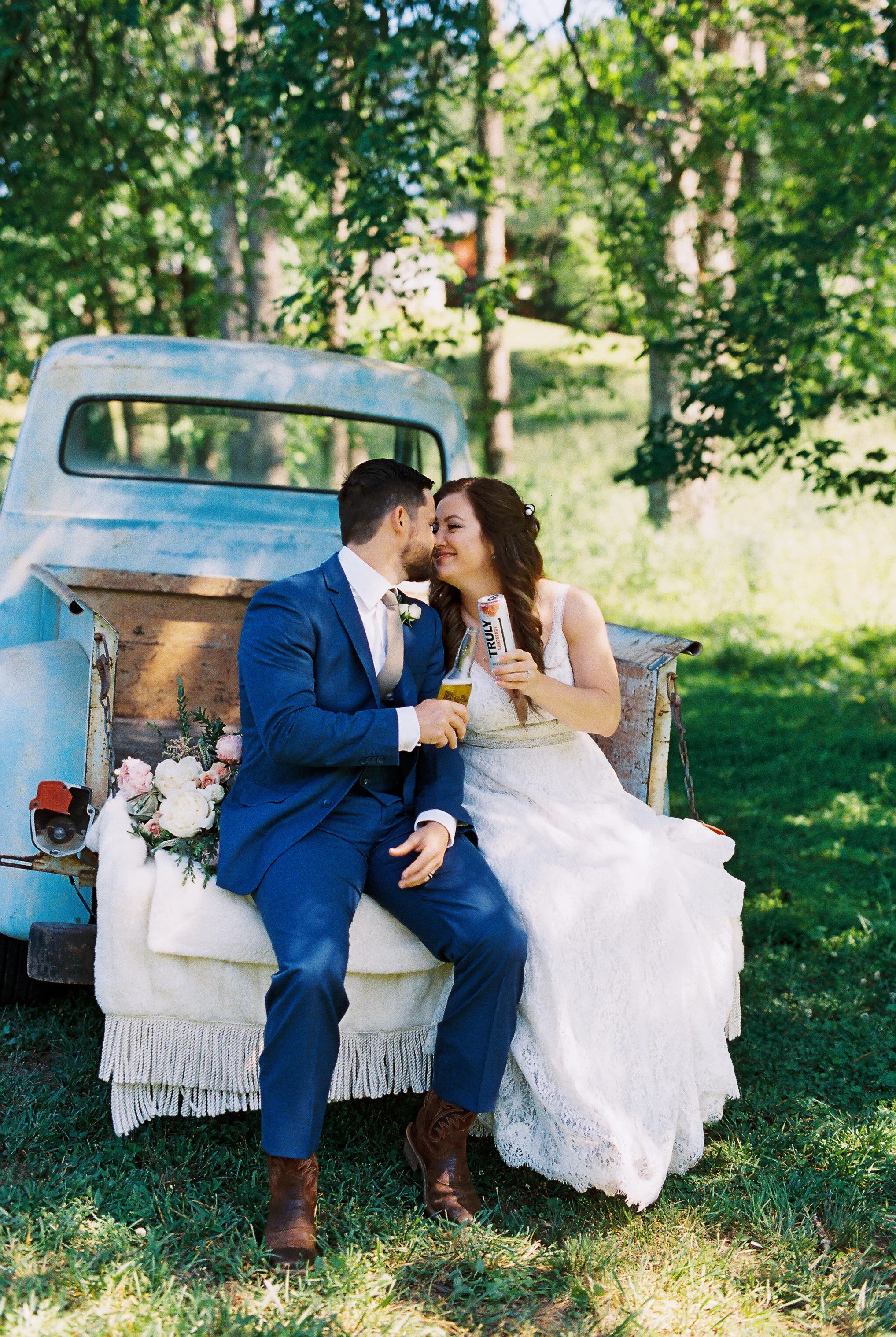 Bride and groom sitting on the vintage truck's tailgate sharing a toast with small drinks, faces close together, bouquet resting beside them