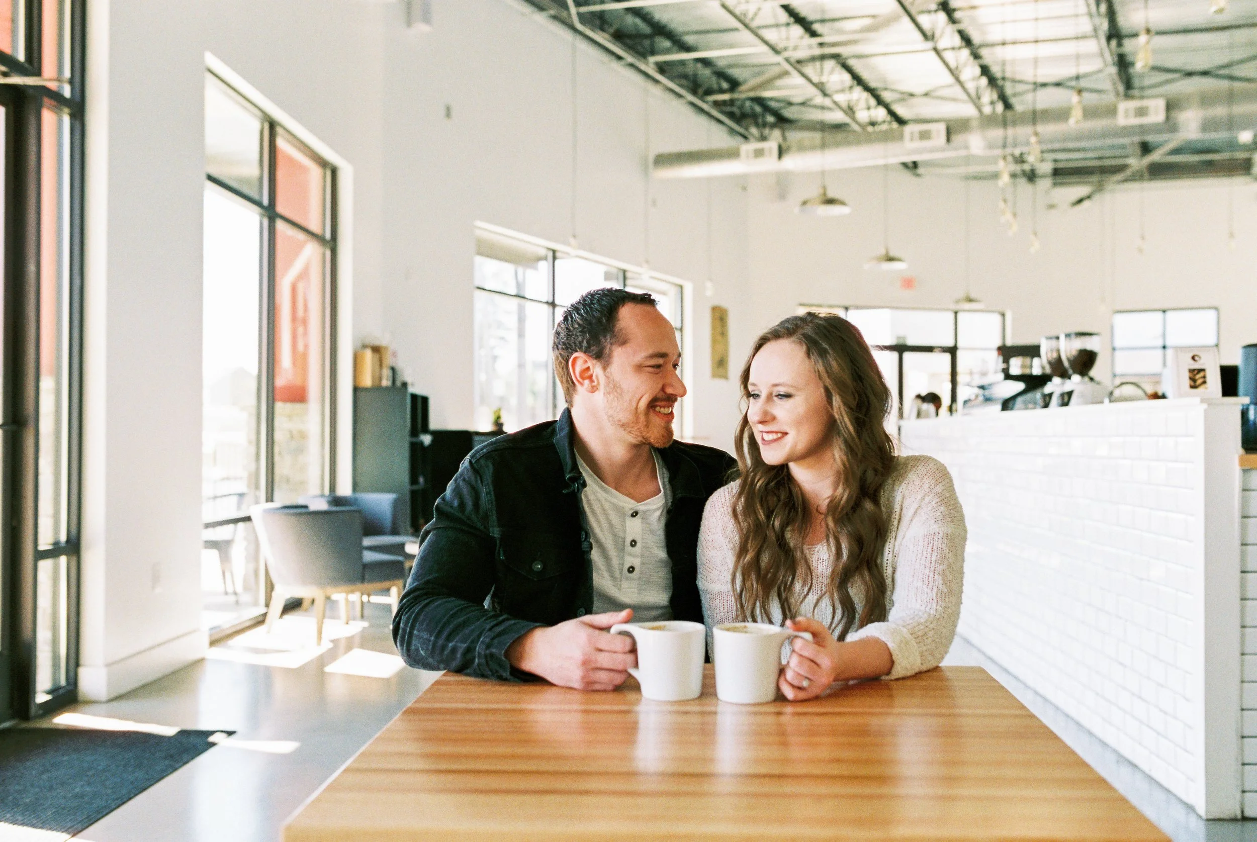 Conner and Maddie smile at each other over coffee mugs at a wide table in a Dallas, Georgia coffee shop.