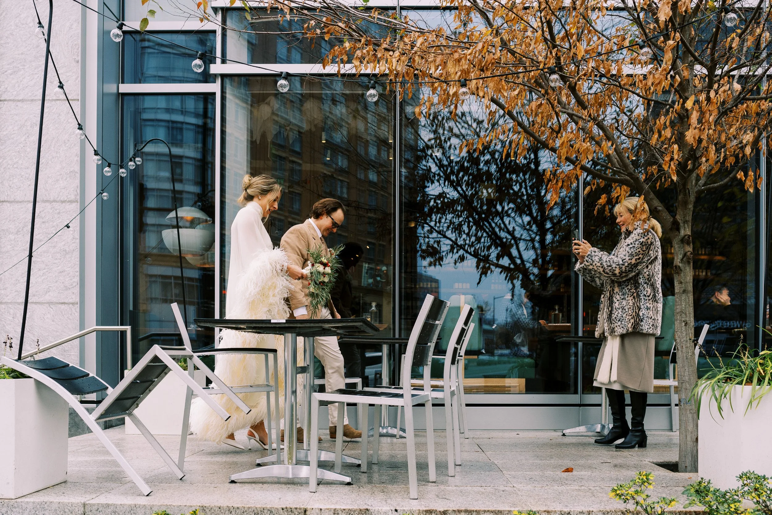 Bride and groom standing at a glass-front restaurant patio with autumn leaves overhead