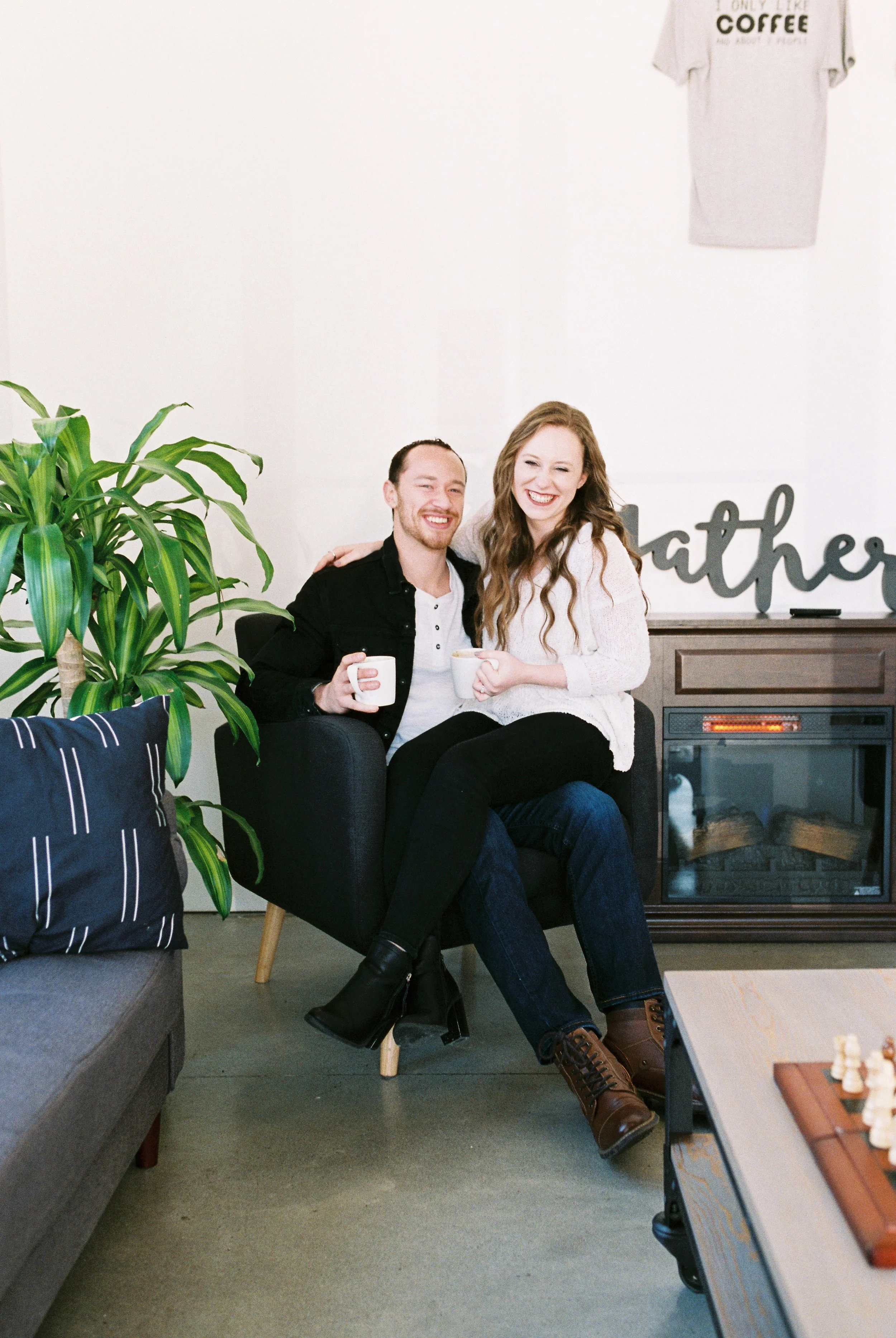 Conner and Maddie beam at the camera holding coffee mugs while sharing an armchair by the fireplace in a Dallas, Georgia coffee shop.