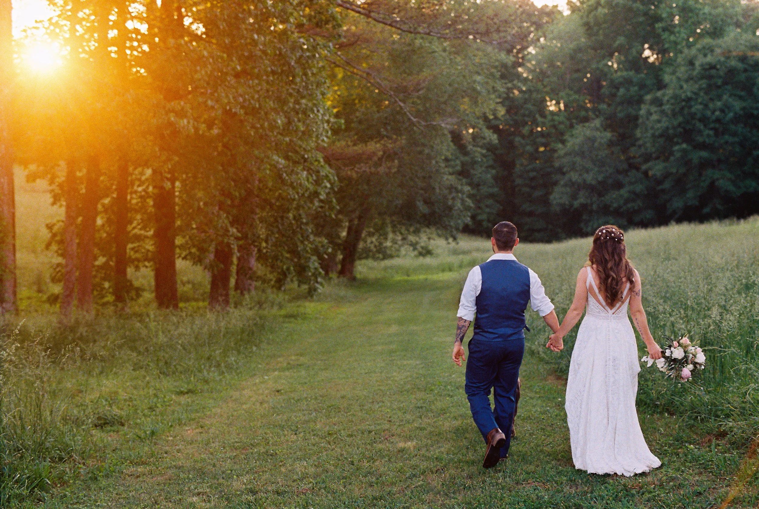Bride and groom walking hand in hand down a grassy path through the trees at golden hour, bride holding her bouquet